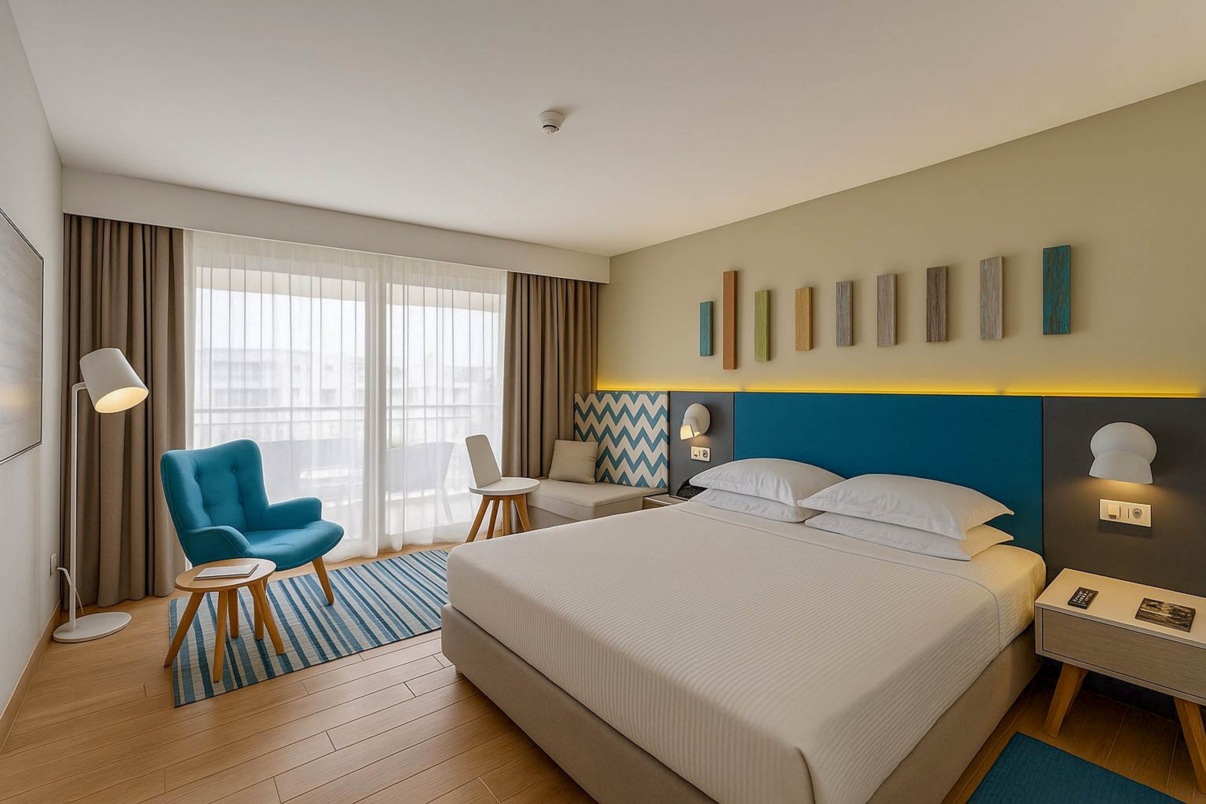 A contemporary hotel room features a neatly made double bed with white linens and a blue upholstered headboard. The space includes a teal armchair, small round tables, and a light-colored sofa near large windows with sheer curtains. Warm wooden flooring, patterned rug, and geometric wall art add visual interest. Soft ambient lighting from bedside lamps and a floor lamp creates a calm, inviting atmosphere.