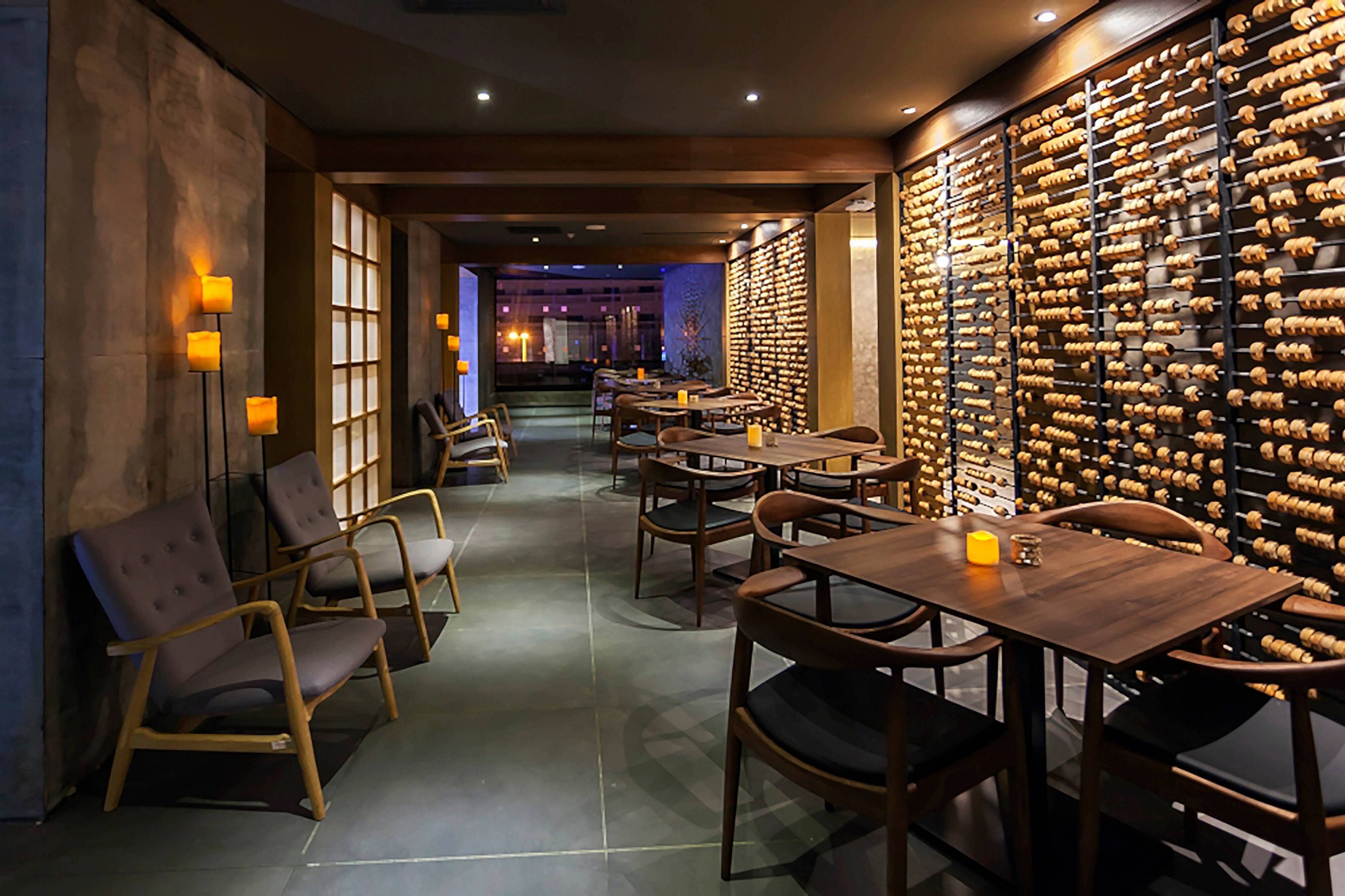 A contemporary wine bar interior features wooden tables and minimalist chairs arranged along a narrow corridor. One wall is lined with an extensive wine rack filled with bottles, creating a focal point for the space. Warm candle-like lights on the tables and wall sconces provide soft, ambient illumination. The overall atmosphere is cozy and sophisticated, ideal for intimate dining or wine tasting.