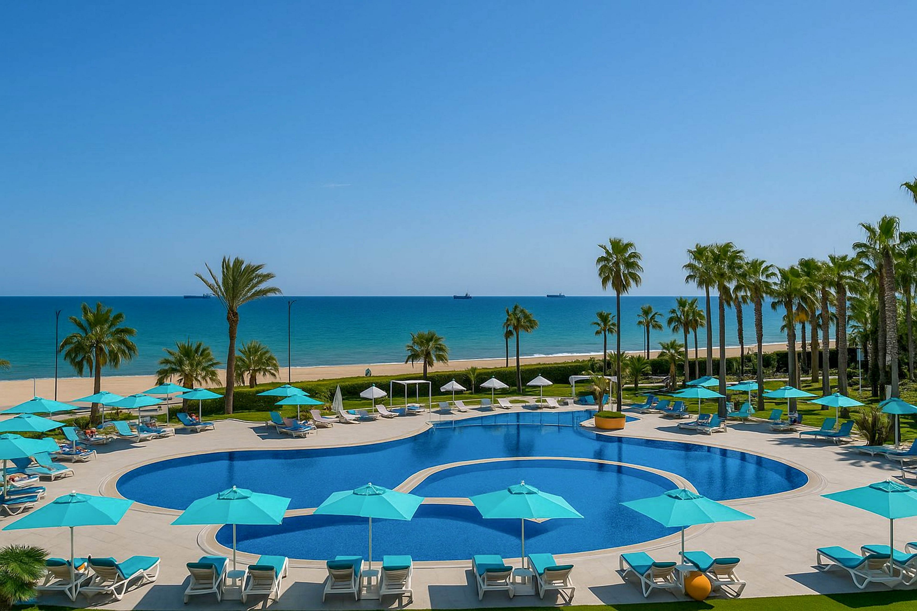 A large resort swimming pool with a distinctive rounded shape sits in the foreground, surrounded by numerous sun loungers and turquoise umbrellas. Tall palm trees line the edge of the pool area, leading the eye toward a sandy beach and calm blue sea. The sky is clear and bright, suggesting a warm, sunny day ideal for relaxation. The overall scene conveys a tranquil, luxurious coastal vacation setting.
