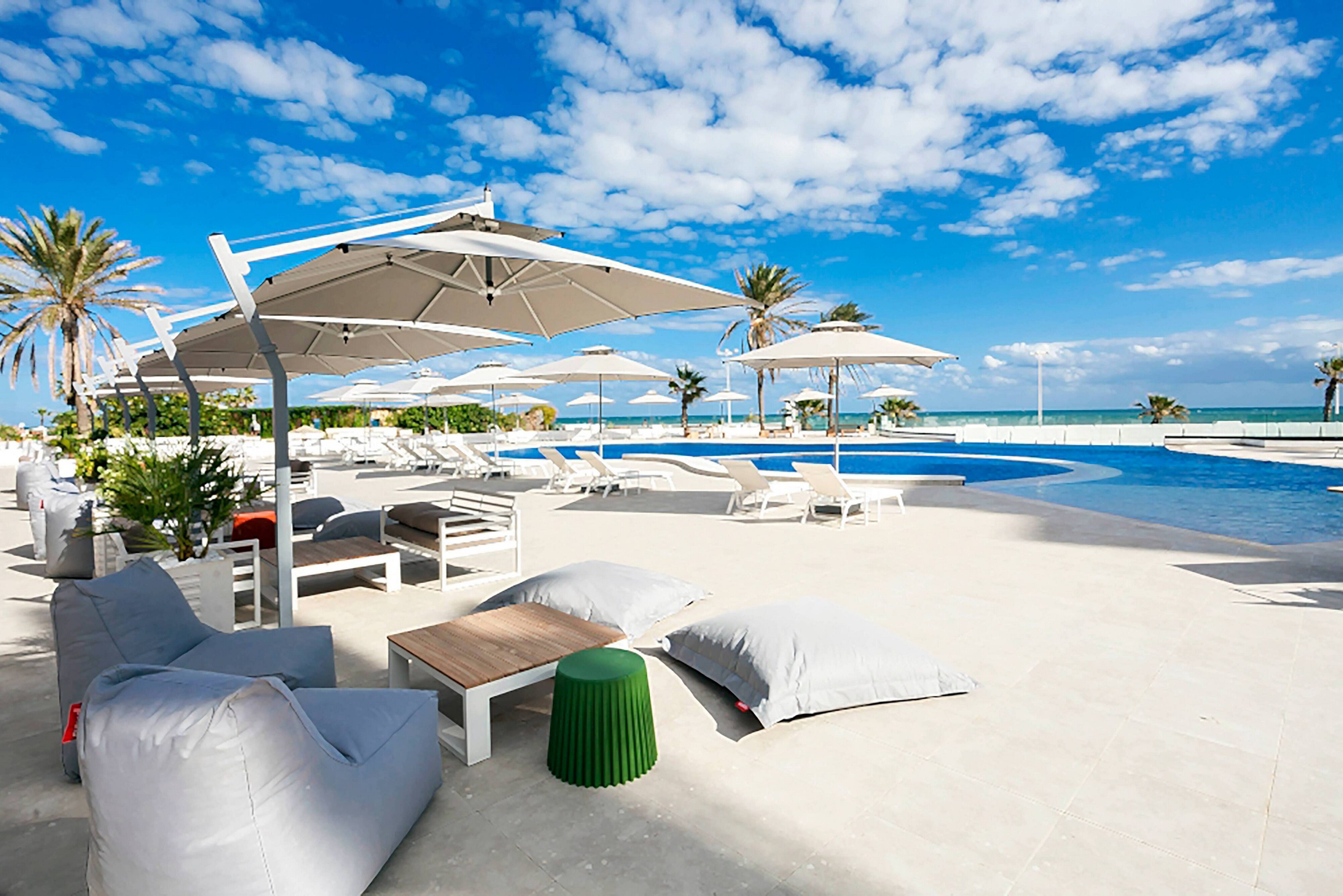 An upscale outdoor pool area overlooks a bright blue sea under a partly cloudy sky. White sun loungers and large umbrellas line the pool deck, creating a resort-style setting. Soft beanbag chairs and low tables provide relaxed seating near the water. Palm trees and lush greenery add a tropical touch to the tranquil, sunlit scene.