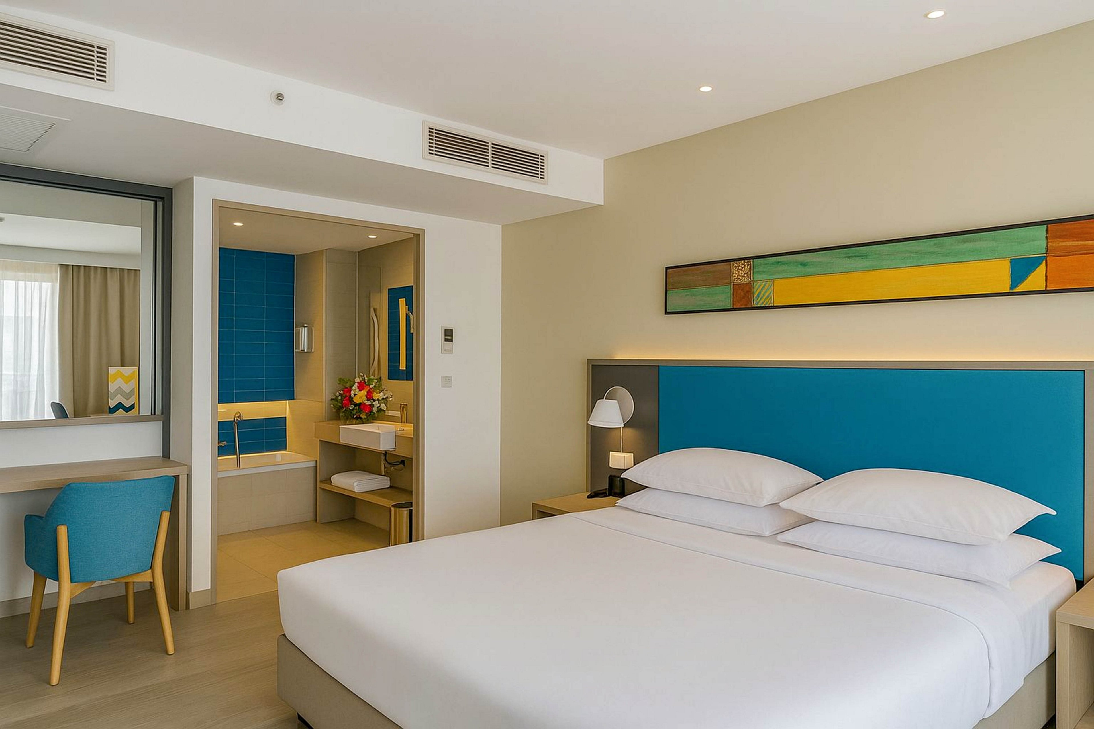 A contemporary hotel room features a neatly made double bed with crisp white linens and a bright blue headboard. An open doorway reveals an ensuite bathroom with blue tiled walls, a bathtub, and a vanity decorated with fresh flowers. A desk and chair sit beside a large mirror, reflecting natural light from the window and sheer curtains. Minimalist decor and warm lighting create a clean, inviting atmosphere suitable for travelers.