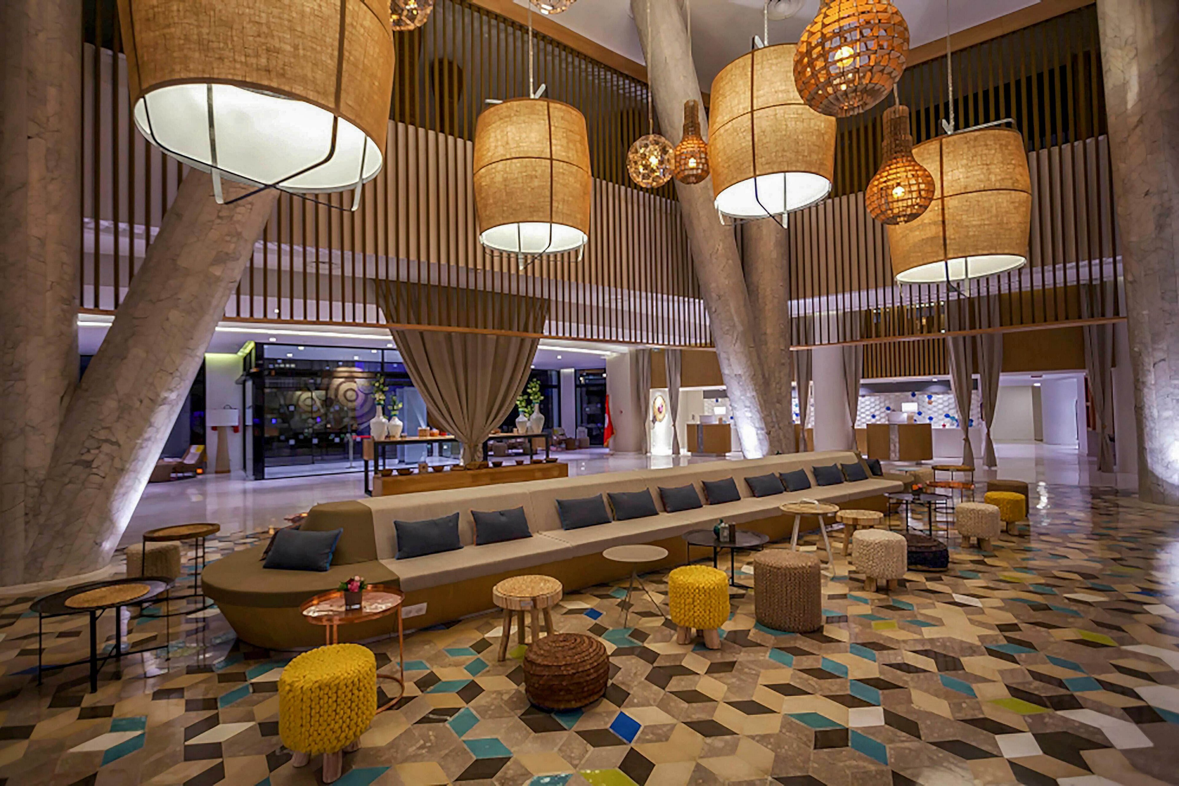 Spacious contemporary hotel lobby featuring a long central sofa and scattered stools. Large woven pendant lights hang from the ceiling, casting warm light over the seating area. The floor is covered with colorful geometric tiles in earth tones and blues. In the background, reception and lounge areas are partially visible behind tall curtains and vertical wooden slats.