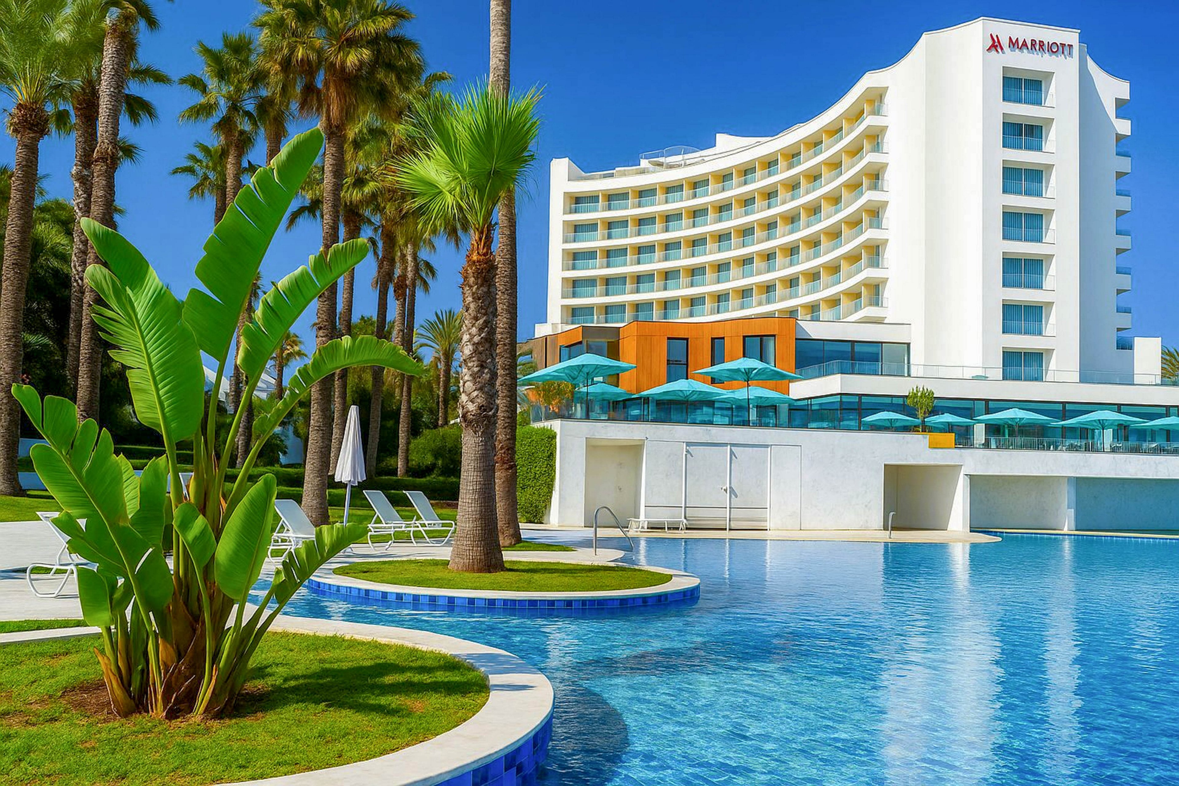 A contemporary Marriott hotel rises behind a large, inviting outdoor swimming pool. Tall palm trees and lush tropical plants frame the clear blue water, creating a resort-like atmosphere. Sun loungers and umbrellas line the pool deck, suggesting a relaxing vacation setting. The building’s curved architecture and bright white facade stand out against the vivid blue sky.