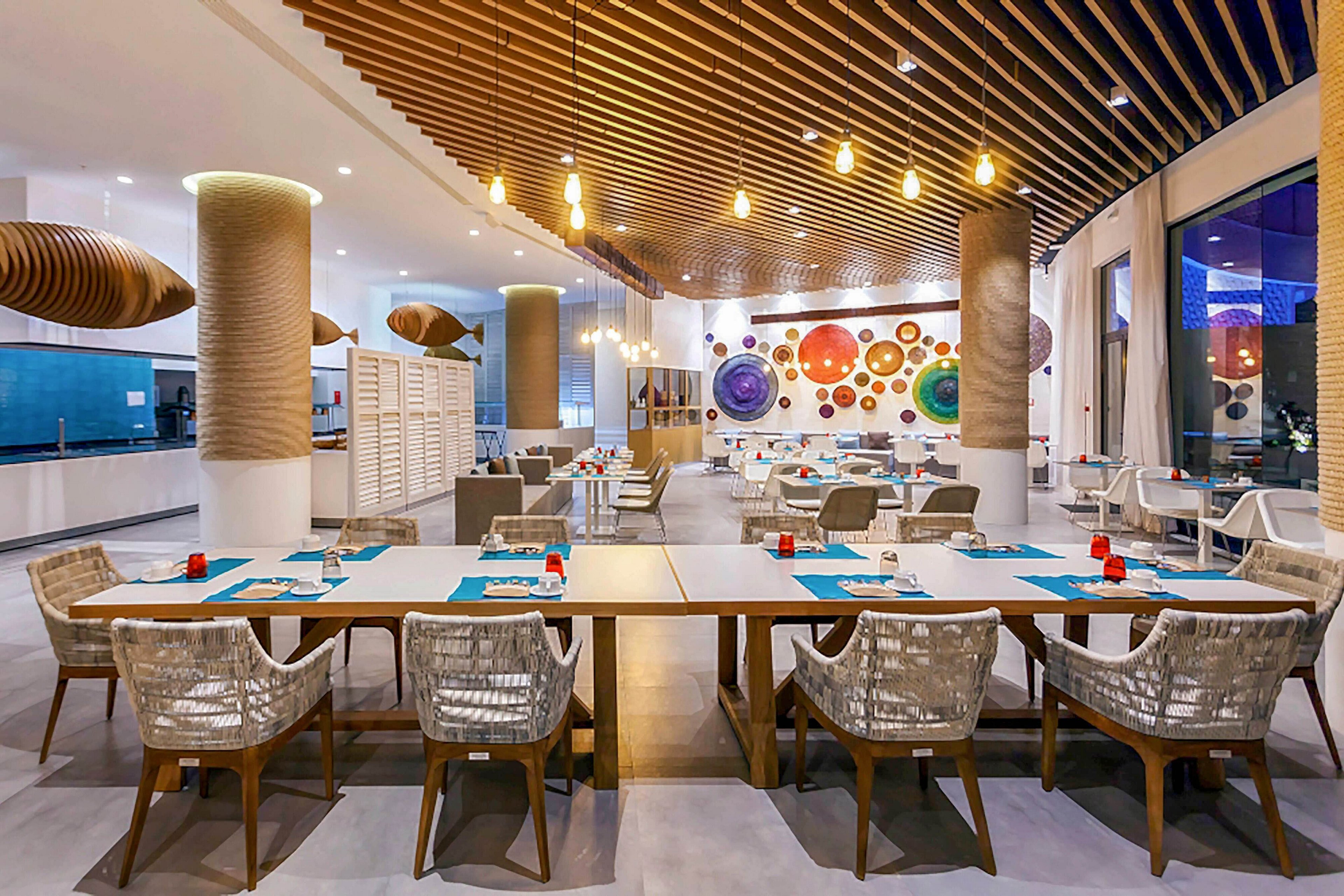 A contemporary hotel restaurant features neatly arranged tables and wicker-style chairs under a wooden slatted ceiling. Warm pendant lights hang above the dining area, creating a cozy yet modern ambiance. The back wall is decorated with large, colorful circular art pieces that add a playful touch to the space. Floor-to-ceiling windows and bright interior lighting make the room feel open and inviting.