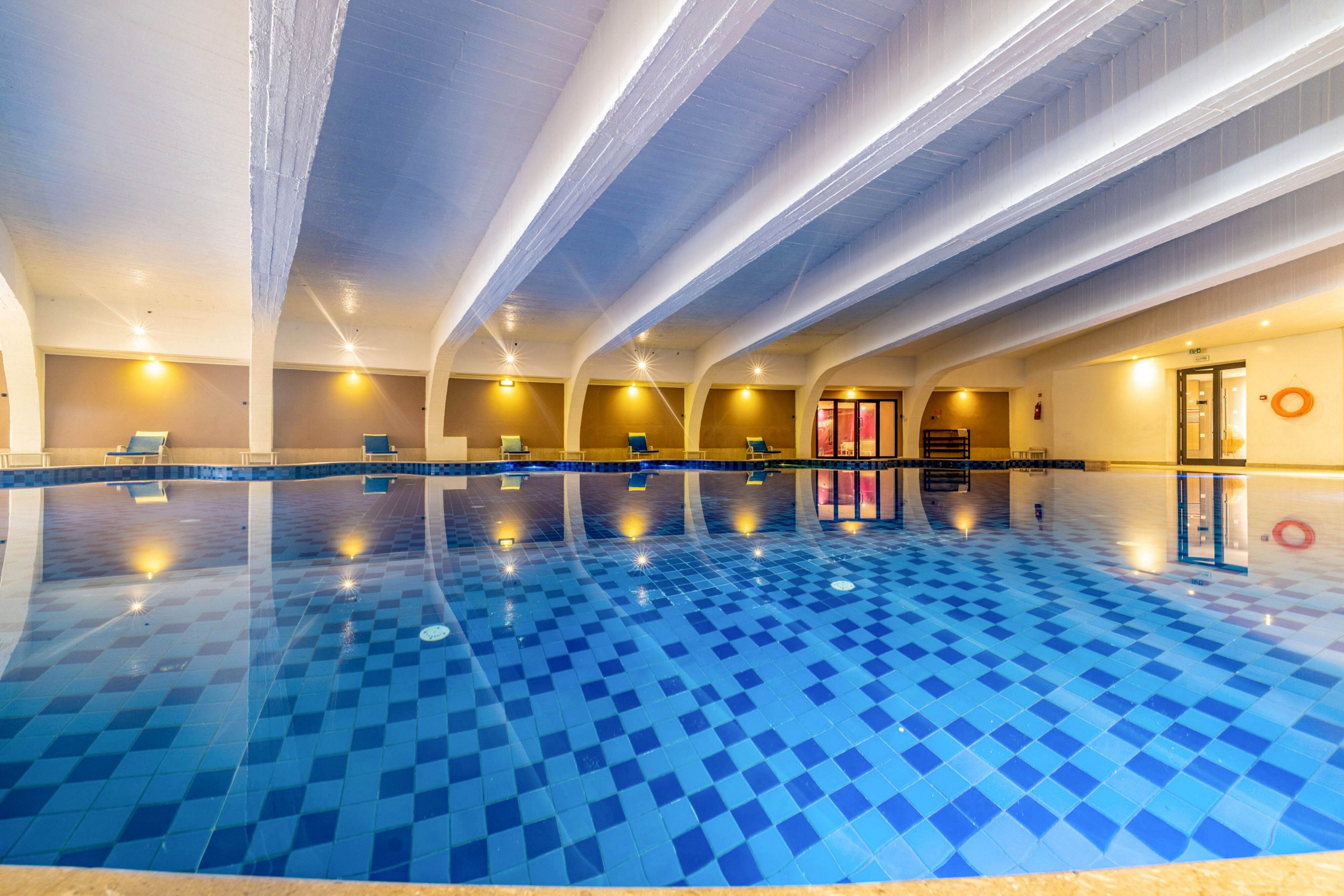 A large indoor swimming pool dominates the scene, featuring a patterned blue tiled floor. The water reflects the ceiling beams and warm wall lights, creating a sense of symmetry. Lounge chairs line the far side of the pool, suggesting a relaxing recreational area. The overall atmosphere is calm and inviting, suitable for leisure or hotel amenities.
