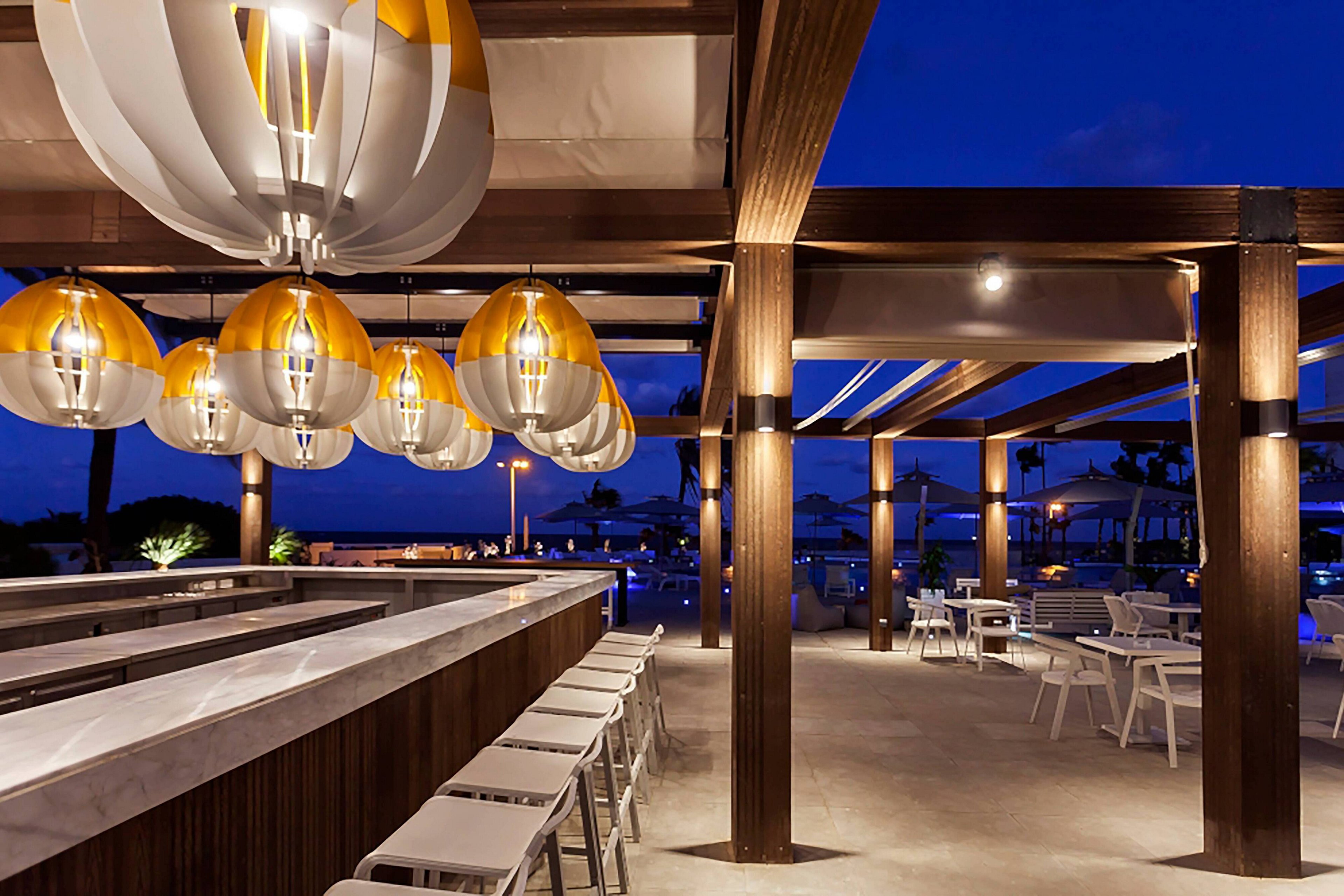 A contemporary open-air bar is shown under a wooden pergola at dusk. Large sculptural pendant lights hang above a sleek marble counter with white bar stools. In the background, poolside seating and palm trees are softly illuminated against a deep blue evening sky. The scene suggests a relaxed vacation atmosphere in a stylish resort setting.