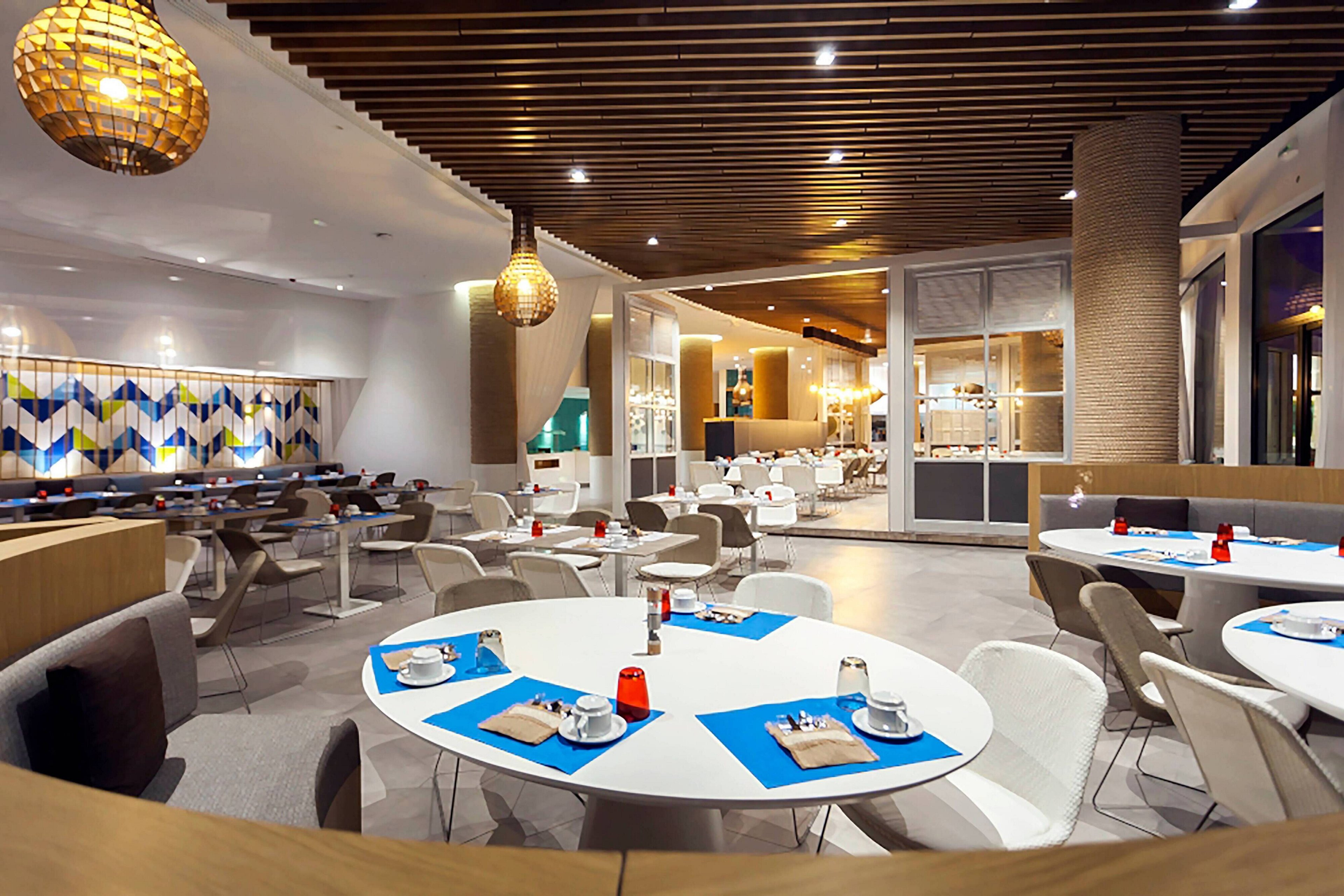 A contemporary restaurant interior features round white tables set with blue placemats and neatly arranged tableware. The spacious dining area is empty, with white and gray chairs surrounding the tables. Warm pendant lights and a wooden slatted ceiling create a cozy yet modern atmosphere. Large windows and patterned wall panels add brightness and visual interest to the space.