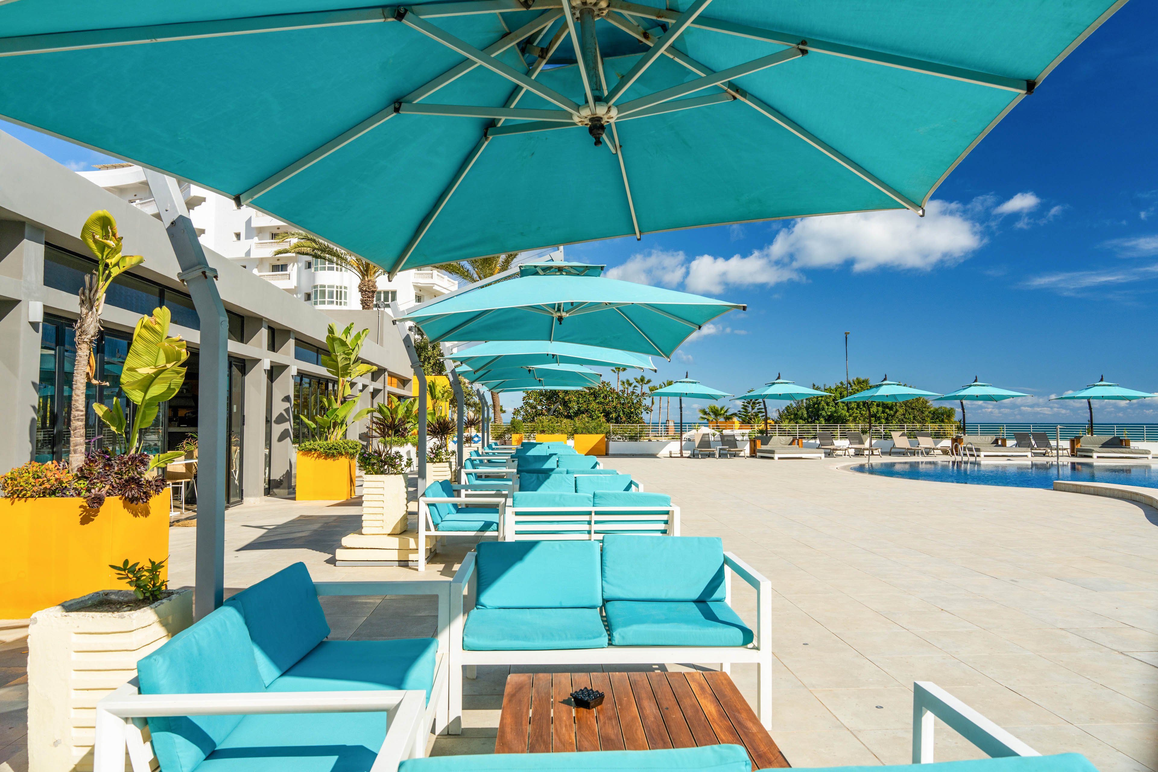 An outdoor resort terrace features turquoise lounge chairs and matching umbrellas beside a swimming pool. Bright yellow planters with tropical plants line the edge of the building, adding contrast to the clean white architecture. The sky is clear and blue, suggesting a sunny, warm day ideal for relaxing by the water. The scene conveys a modern, upscale vacation atmosphere with vibrant colors and open space.