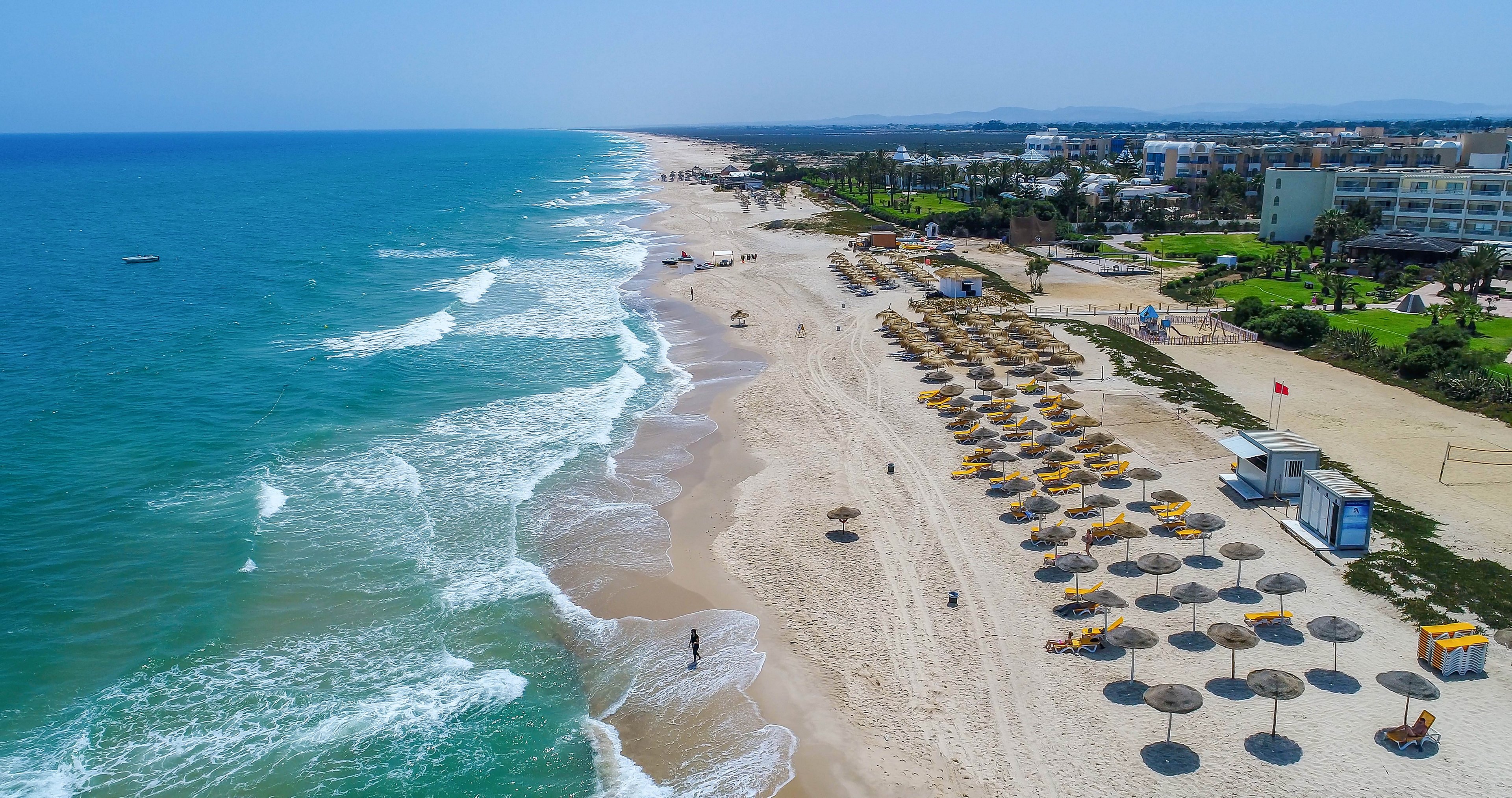 A wide sandy beach stretches along a turquoise sea viewed from above. Neatly arranged sun loungers and umbrellas line the shore near a coastal resort complex. Gentle waves roll onto the sand where a few people walk by the water. The scene suggests a warm, sunny day at a popular seaside holiday destination.