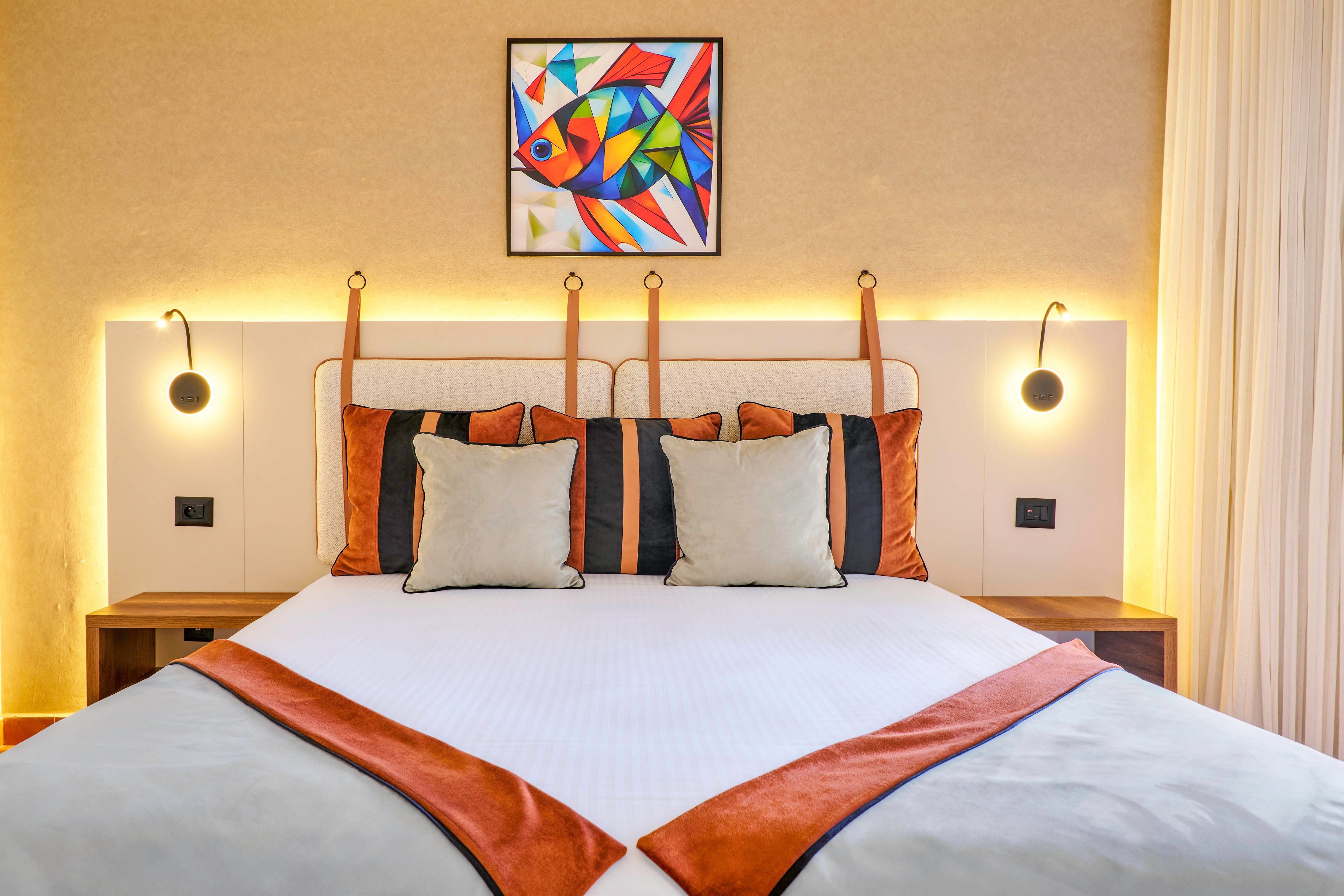 A neatly made double bed is centered in a modern hotel-style bedroom. The bed features white linens with rust-colored accents and matching decorative pillows. Warm backlighting highlights the upholstered headboard and two wall-mounted reading lights on either side. Above the bed hangs a vibrant geometric painting of a fish, adding a colorful focal point to the neutral-toned room.