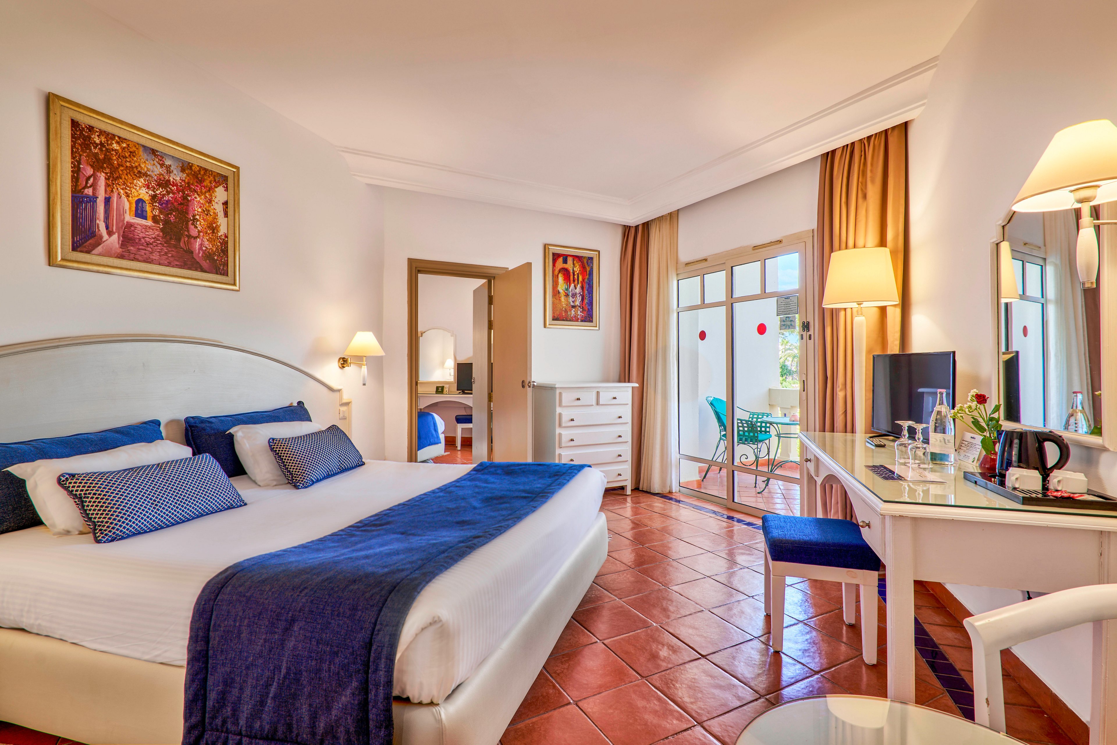 Spacious hotel room featuring a large neatly made bed with blue and white bedding. The space includes a desk with a television, kettle, and chair, as well as a dresser and wall art. Large windows with curtains open onto a terrace with outdoor seating and greenery visible outside. Warm lighting and terracotta floor tiles create a cozy yet airy atmosphere suitable for vacation or business travel.