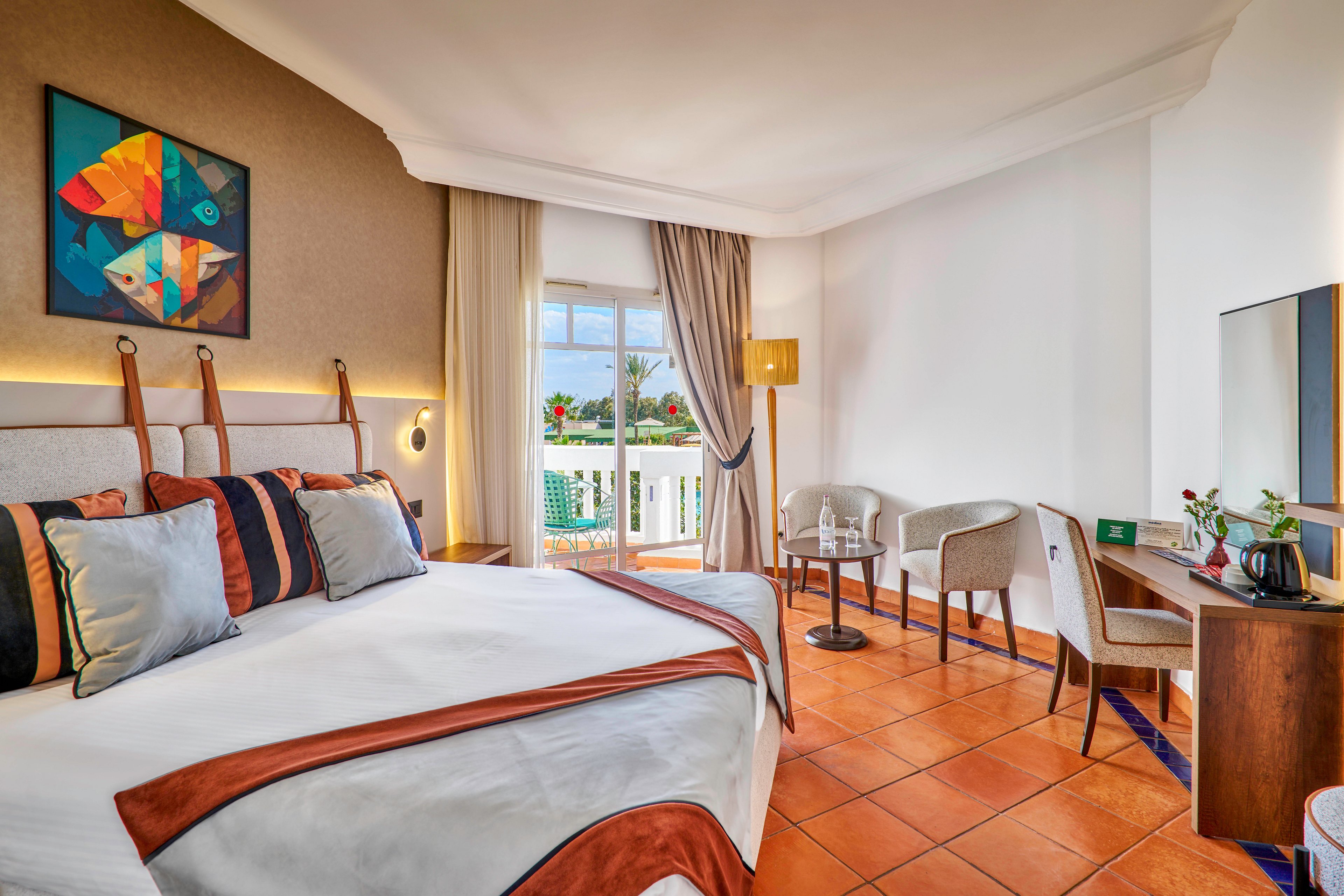 Well-appointed hotel room featuring a large neatly made bed with decorative pillows and throws. The space includes a small seating area with two chairs and a round table near a floor lamp. A desk with a mirror, chair, and small decor items sits opposite the bed. Warm terracotta floor tiles and a colorful fish artwork add vibrancy, while a balcony door opens to a sunny outdoor view with greenery and buildings.