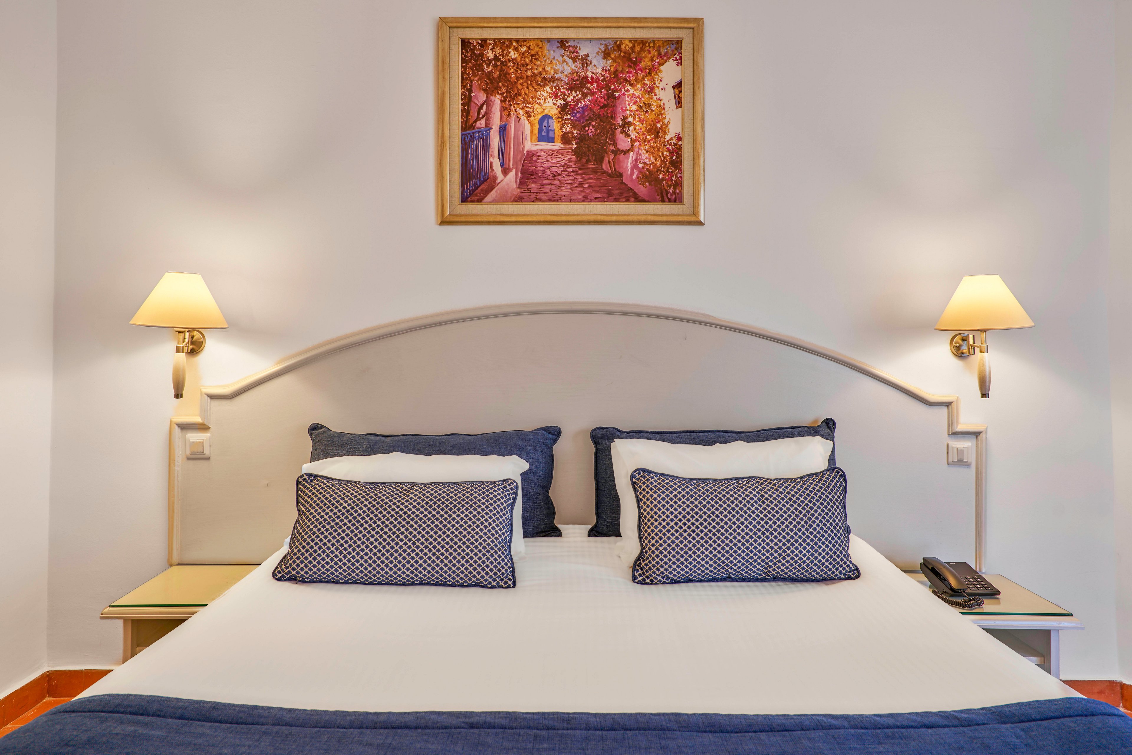 A neatly made double bed with white linens and blue accents is centered against a light-colored wall. Two matching wall-mounted lamps flank the bed, casting a warm, inviting glow. Decorative pillows and a blue bed runner add contrast and texture to the scene. A colorful framed painting hangs above the headboard, completing the simple, cozy hotel room decor.