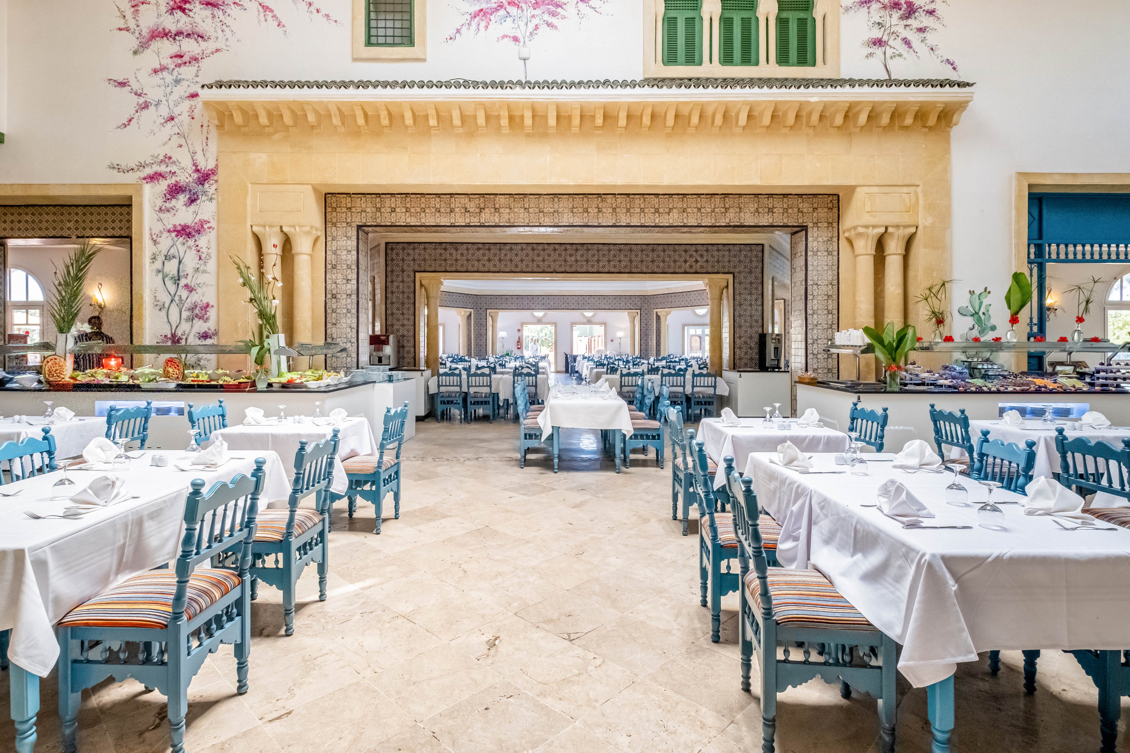 Spacious restaurant interior with neatly arranged tables and blue wooden chairs is shown in a bright, airy setting. White tablecloths and folded napkins create a formal dining atmosphere, while colorful striped chair cushions add warmth. Decorative arches, floral wall art, and traditional architectural details frame the view through multiple dining areas. Buffet counters with fruits and dishes line the sides, suggesting a hotel or resort dining experience.