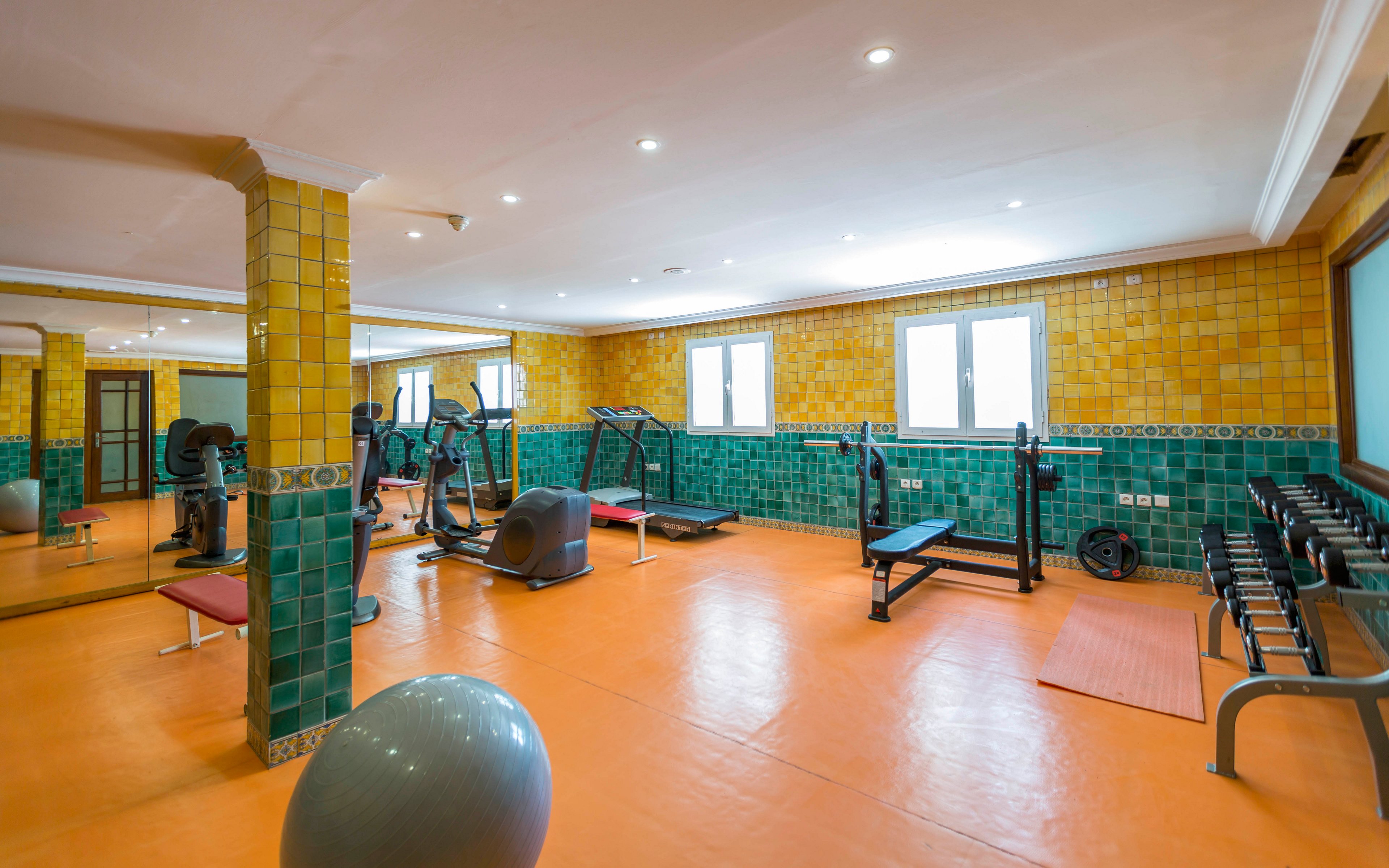 A spacious indoor gym features various fitness machines and free weights arranged neatly along the walls. The room has orange flooring with green and yellow tiled walls, creating a colorful and clean environment. Large windows allow natural light to brighten the space, complemented by recessed ceiling lights. A large mirror, exercise ball, benches, and dumbbell rack emphasize the room’s focus on strength and cardio training.