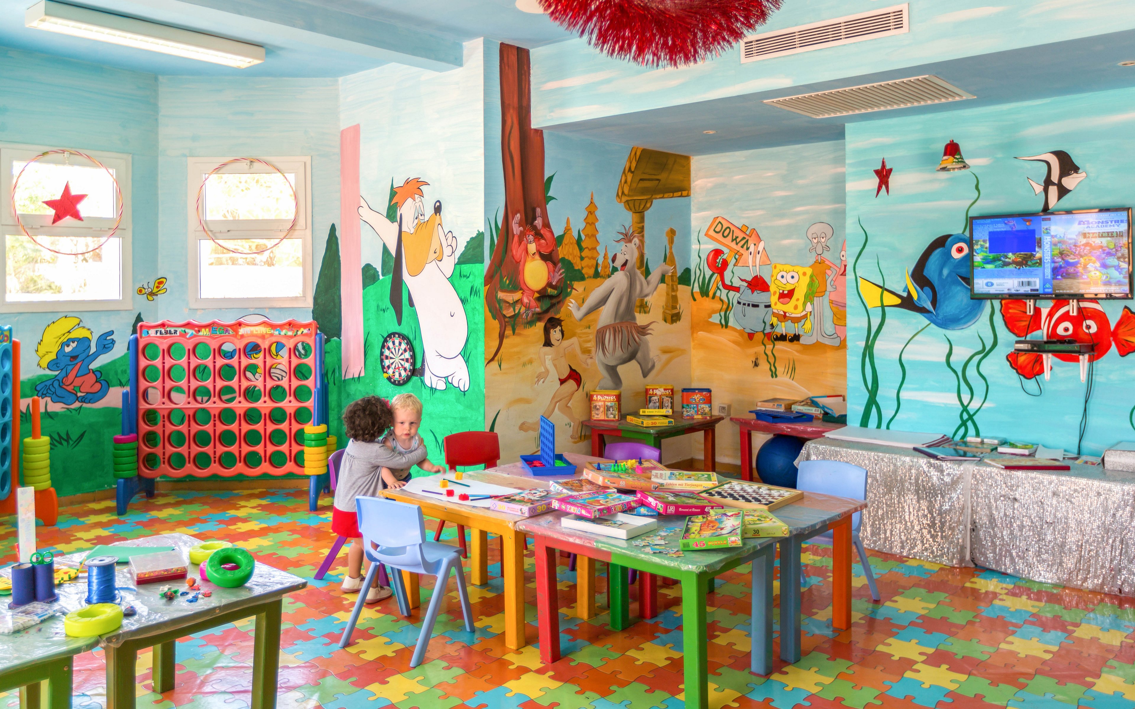 A bright children’s playroom features two young kids seated at a table surrounded by toys and art supplies. The room has vividly painted cartoon murals covering the walls, depicting underwater scenes and animated characters. Colorful interlocking floor tiles, small chairs, and multiple activity tables create a playful learning environment. A large vertical game board and a wall-mounted television add to the variety of entertainment options in the space.