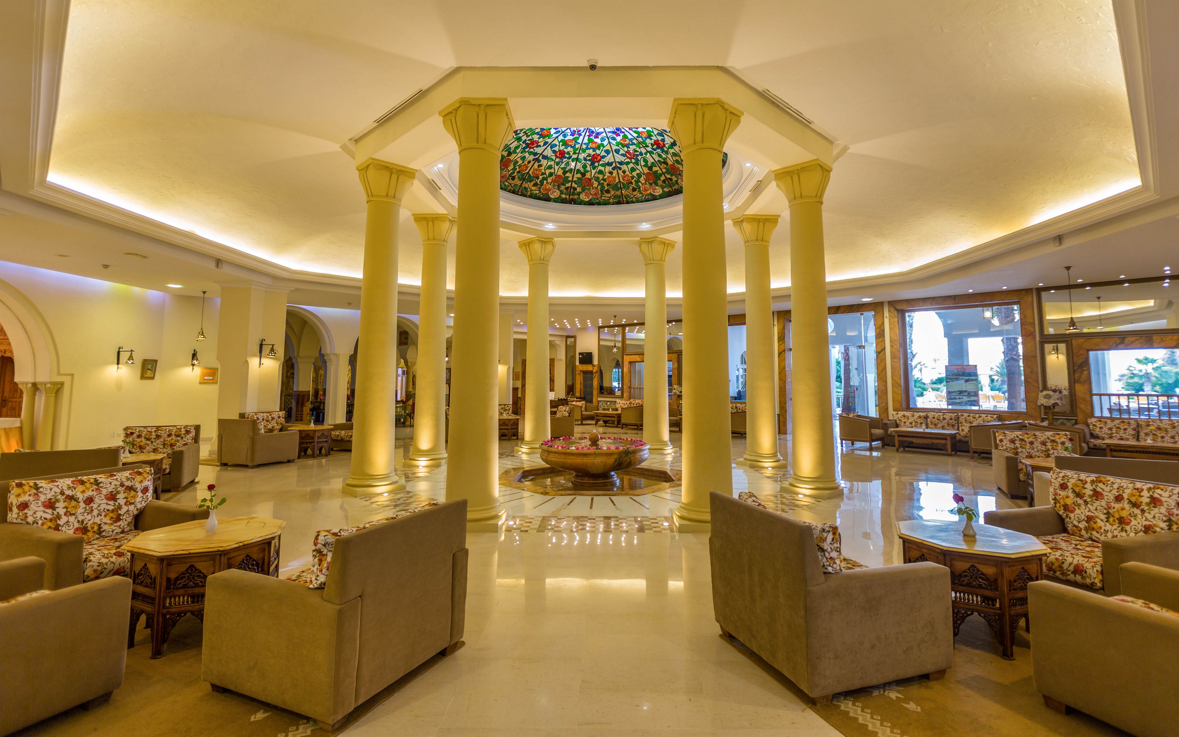 A spacious hotel lobby features tall cream-colored columns arranged in a circular pattern around a central seating area. Warm ambient lighting highlights the polished floor and the decorative stained-glass ceiling above. Comfortable sofas and armchairs with floral upholstery are arranged in small conversation groups throughout the space. Large windows and archways in the background suggest access to other parts of the hotel and views outside.