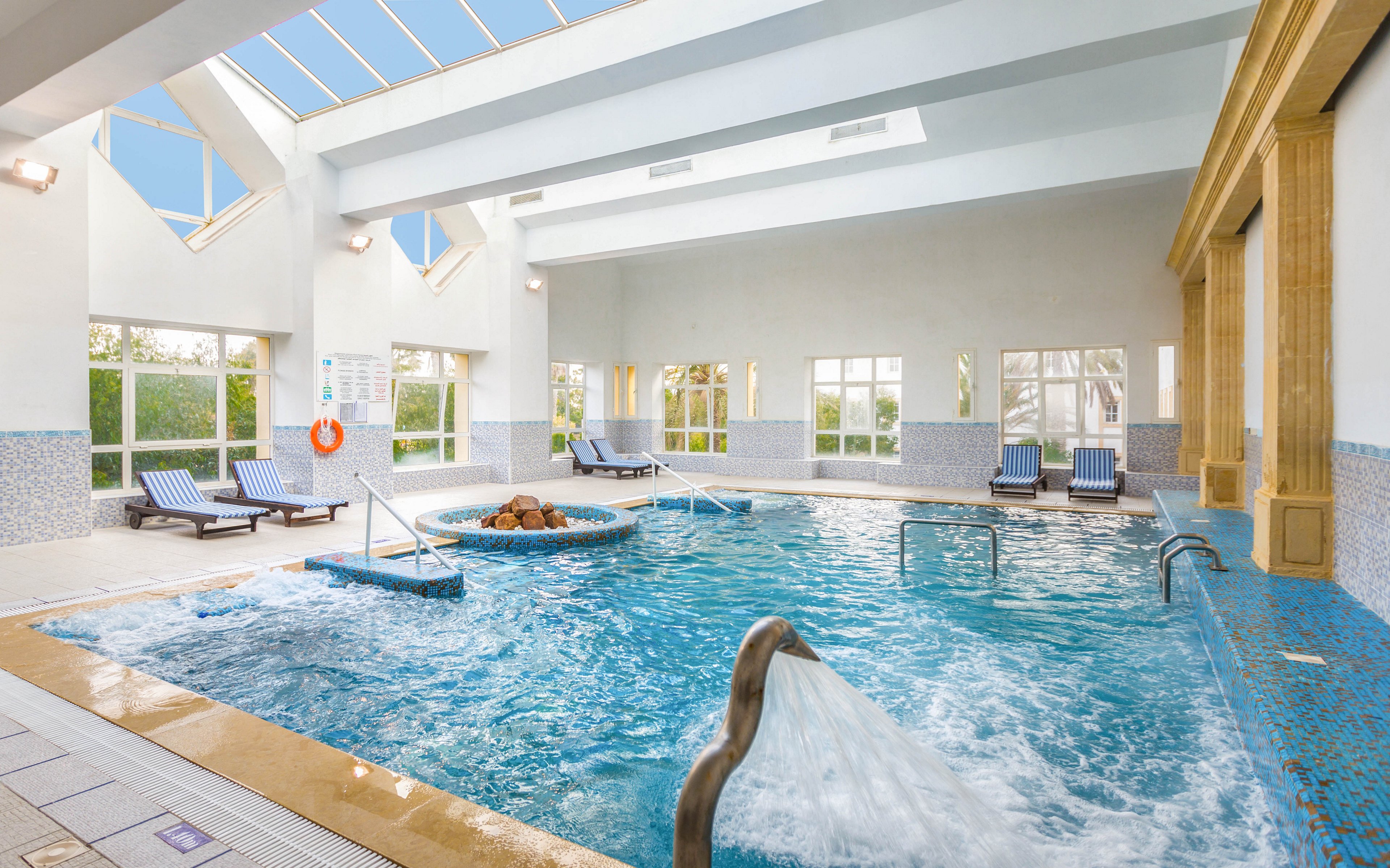 A spacious indoor swimming pool features clear blue water and a central circular spa area with bubbling jets. Large skylights in the ceiling and tall windows along the walls fill the room with natural light. Several striped lounge chairs and small tables line the poolside, creating a relaxing resort-like atmosphere. The clean architectural lines and light color palette emphasize a calm, modern wellness setting.