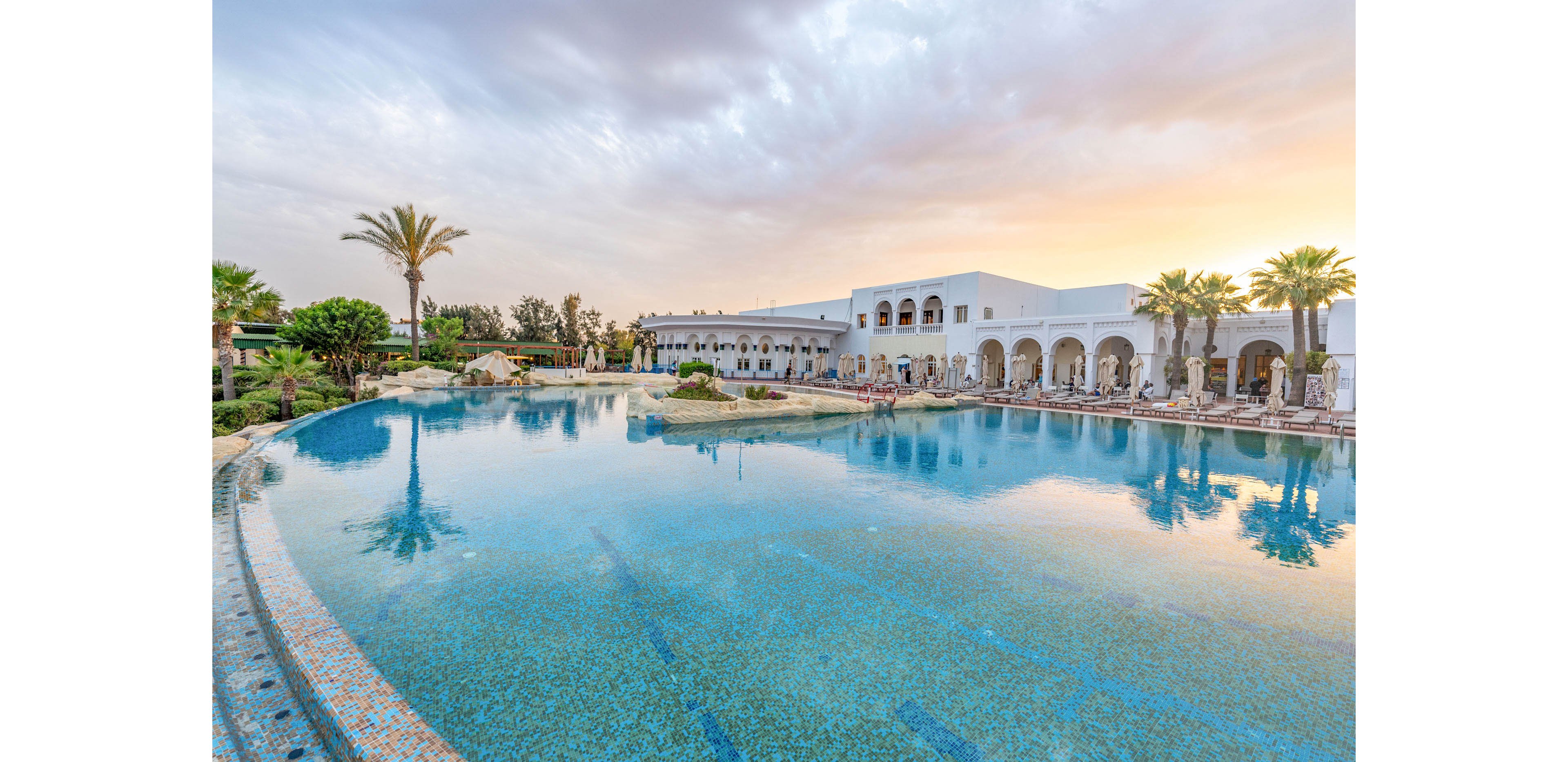 A large, tranquil swimming pool reflects a white resort building with arched colonnades. Palm trees and lush greenery surround the pool, creating a tropical oasis atmosphere. Lounge chairs line the far side of the pool, suggesting a relaxing vacation setting. The sky is partly cloudy with warm sunset tones, adding a serene and inviting mood.