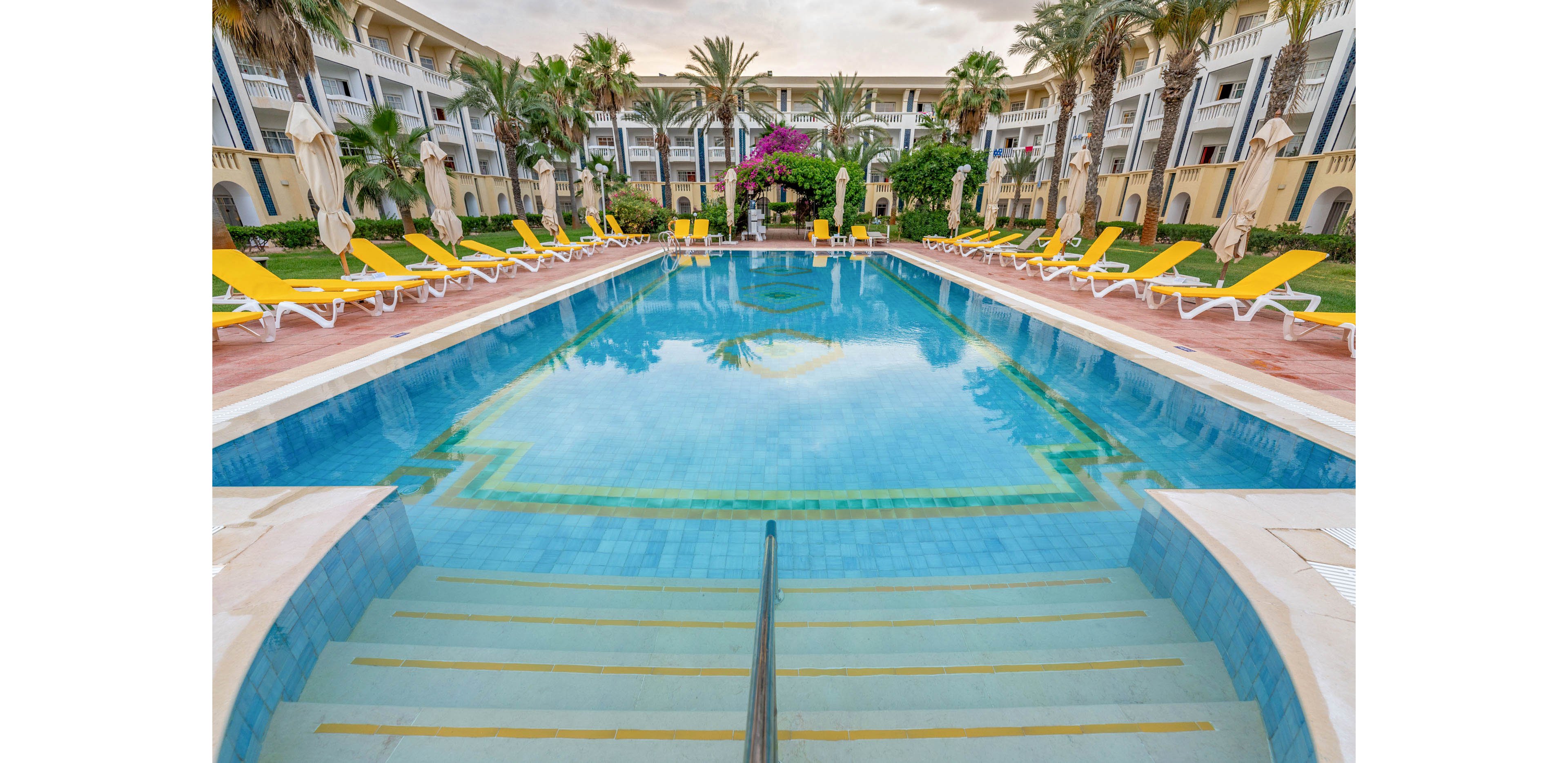 A large resort swimming pool is centered between rows of bright yellow lounge chairs and umbrellas. The pool features wide entry steps and clear blue water reflecting the surrounding architecture. Palm trees and lush greenery frame the scene, with a multi-story hotel building in the background. The setting appears calm and inviting, suggesting a relaxing vacation atmosphere.