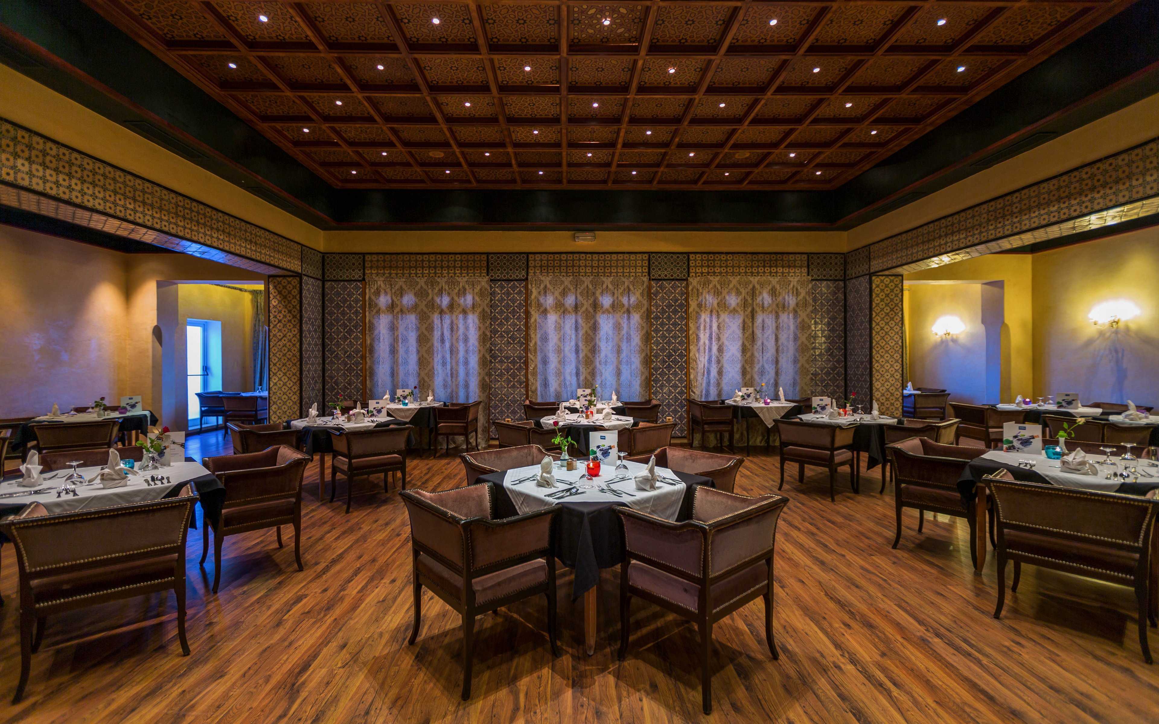 A spacious, upscale restaurant dining room is set with neatly arranged tables and chairs. White tablecloths, folded napkins, and glassware are prepared for guests, but the room is currently empty. Warm yellow lighting and patterned wall panels create a cozy, sophisticated atmosphere. A decorative wooden ceiling and polished wooden floor add to the refined interior design.