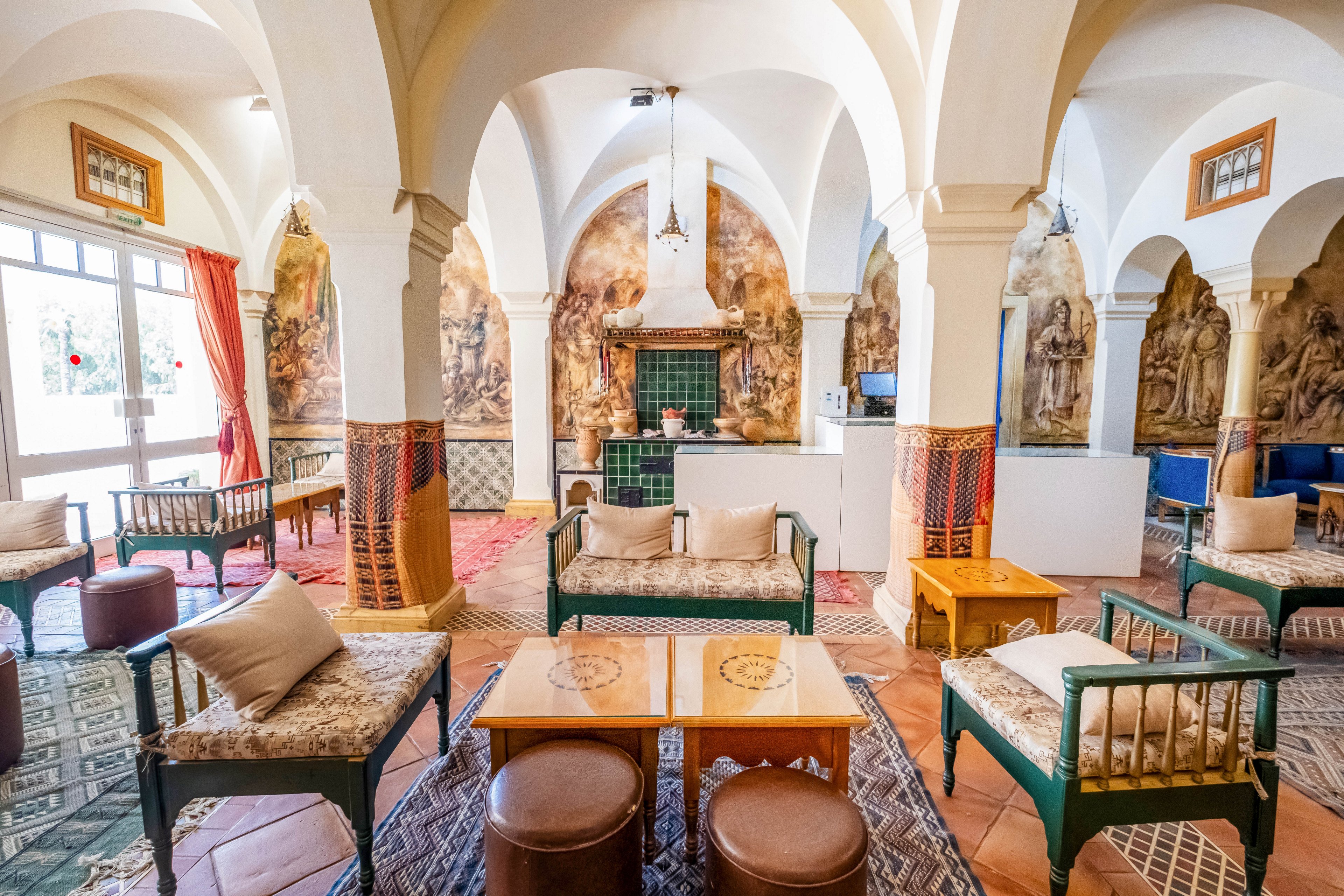 A sunlit lounge features arched ceilings and ornate columns surrounding a cozy seating area. Cushioned benches, armchairs, and small tables are arranged on patterned rugs, creating an inviting social space. Decorative wall art and tiled details add a historic and artistic character to the room. Large windows with curtains allow natural light to flood the interior, highlighting the warm colors and textures.