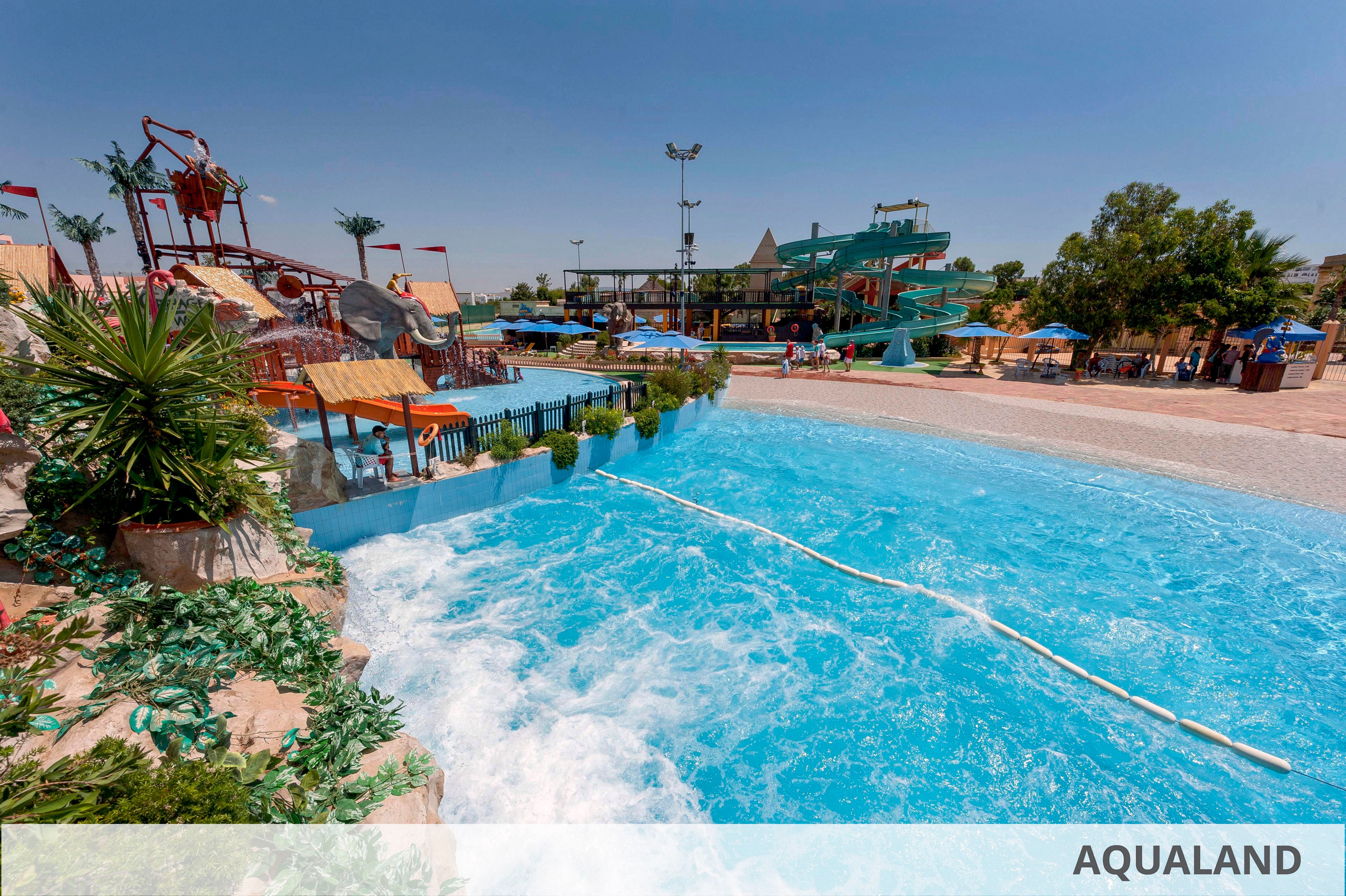 A large outdoor water park features a bright blue wave pool under a clear sunny sky. Multiple waterslides and play structures rise in the background, with people enjoying the attractions and lounging under umbrellas. Green plants and landscaping frame the pool edge, adding a tropical feel to the scene. The word "AQUALAND" appears in text at the bottom right of the image.