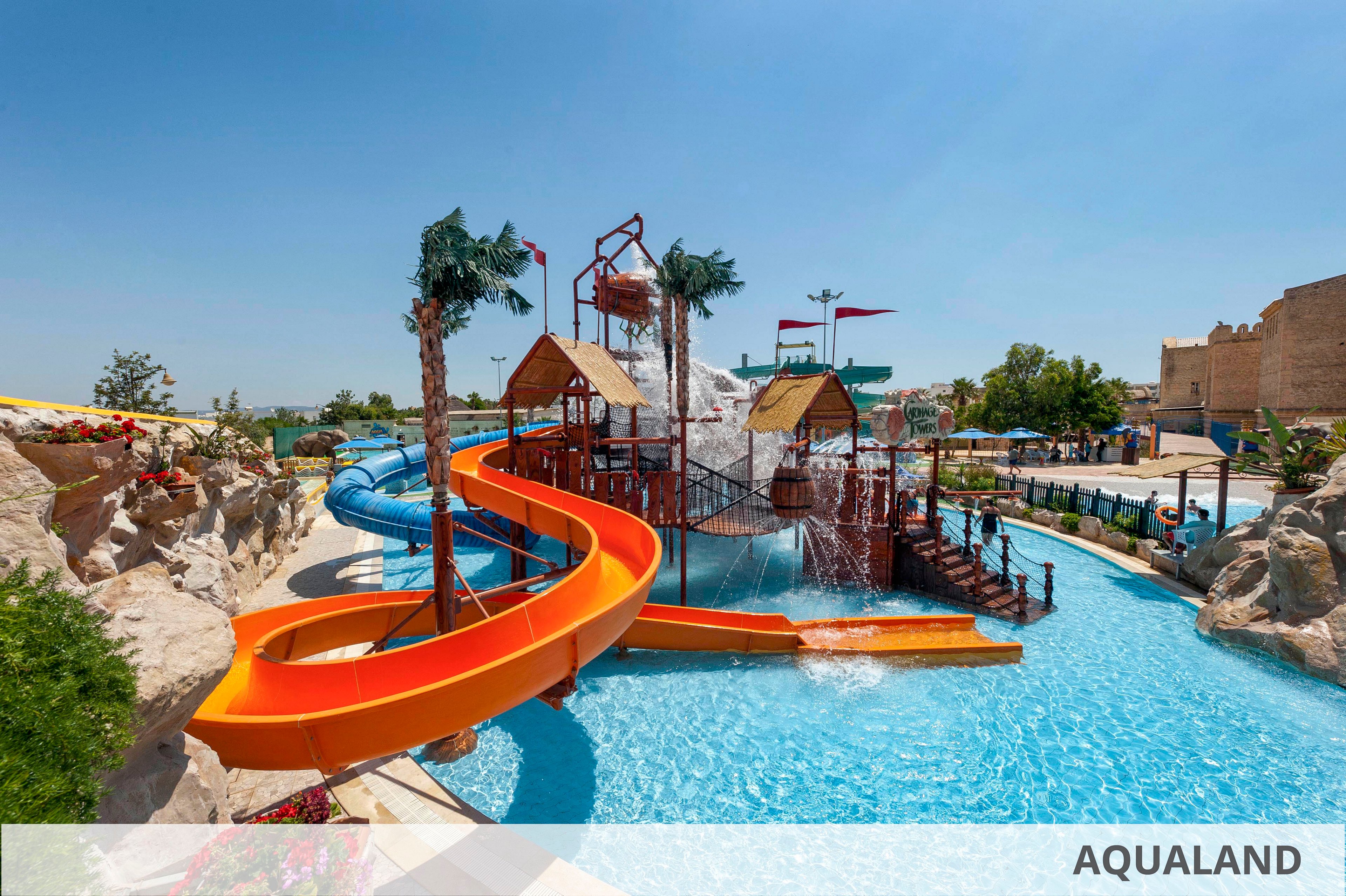 A vibrant outdoor water park features multiple twisting slides and splash structures surrounded by clear blue pools. Orange and blue water slides curve around artificial rock formations and palm trees under a bright sunny sky. Wooden platforms, water buckets, and play towers create an adventurous setting for families and children. The word "AQUALAND" appears in the lower right corner, indicating the name of the attraction.