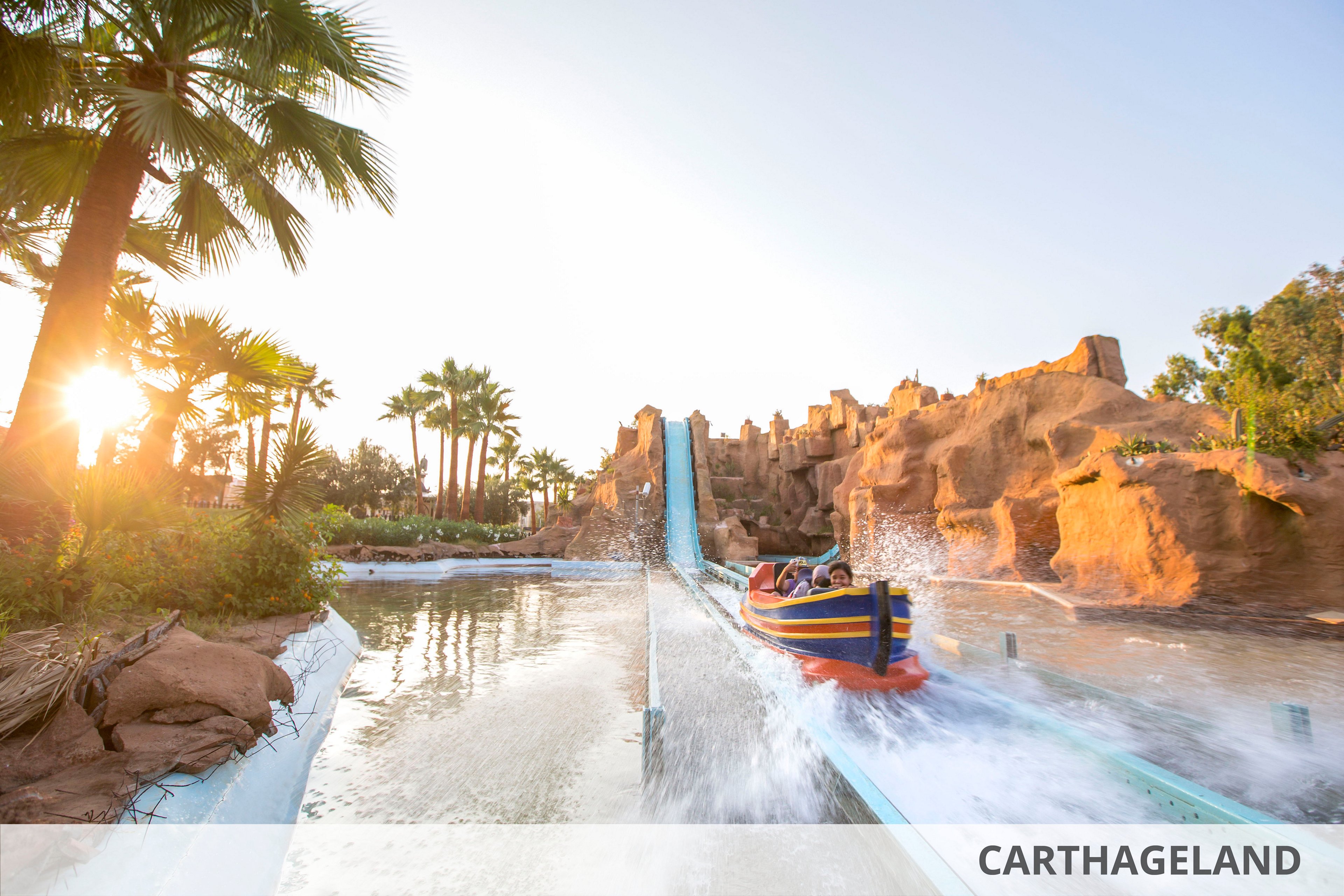 A colorful boat plunges down a steep water ride at Carthageland amusement park. The attraction is surrounded by rocky structures, palm trees, and a bright sunset sky. Visitors seated in the boat create a dramatic splash as they speed along the flume. The scene conveys excitement and fun in a warm, outdoor resort setting.