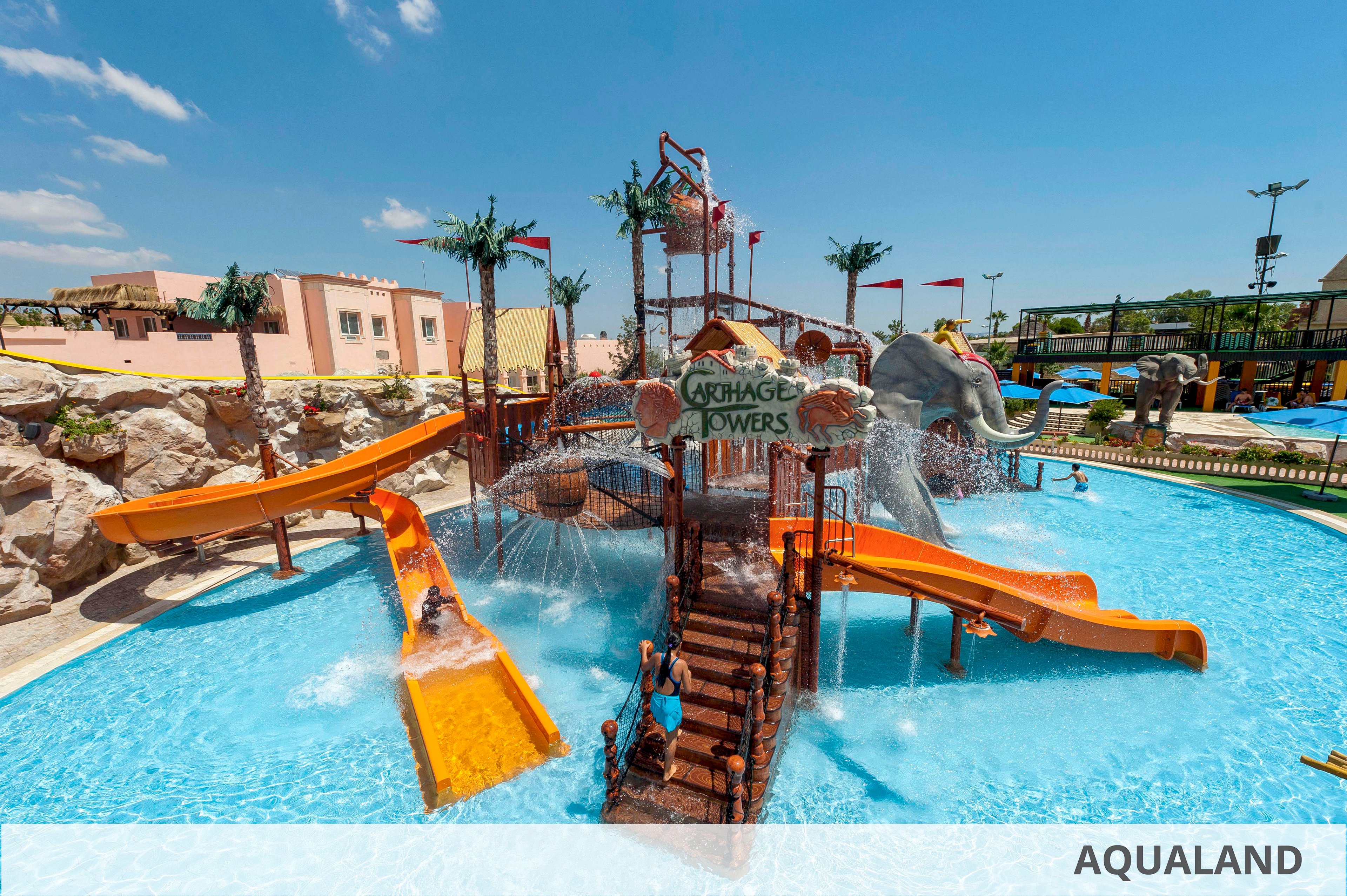 An outdoor water playground features multiple orange slides and a central climbing structure labeled "AQUA GAME TOWERS." Children play in the bright blue pool as water sprays from various fountains and overhead buckets. Palm tree decorations and a large elephant figure enhance the fun, tropical theme. The scene is set under a clear sunny sky with surrounding resort buildings and walkways visible.