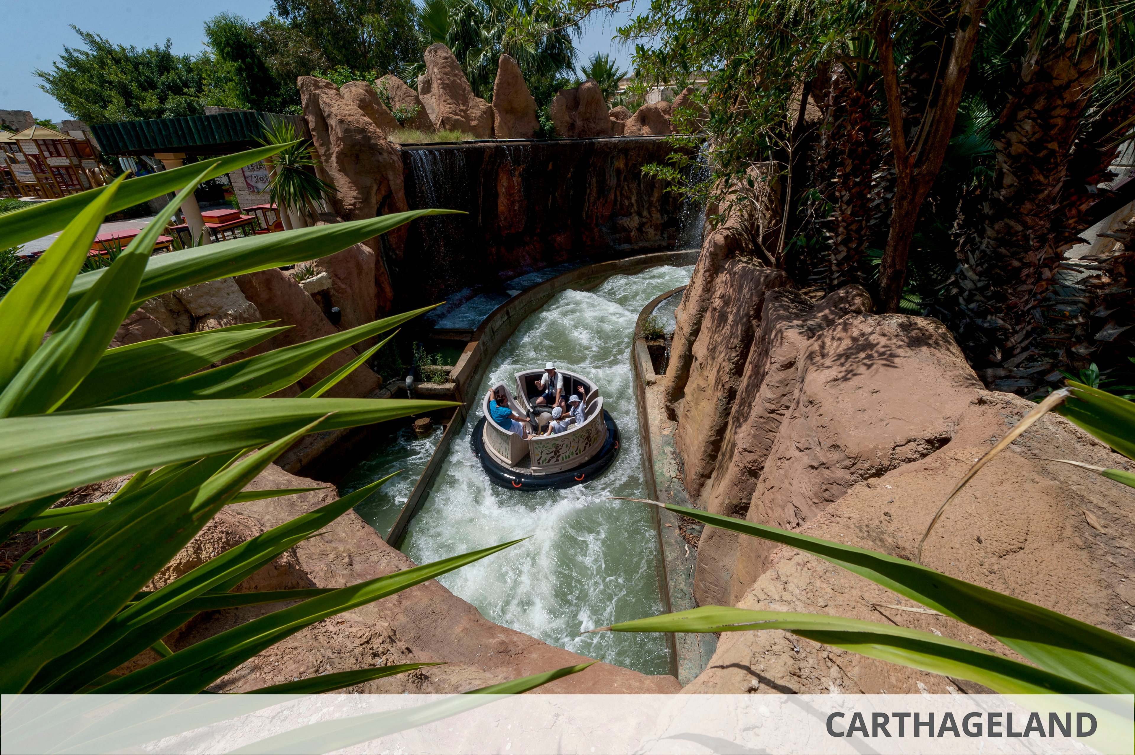 A circular raft carrying several people navigates a fast-moving water channel between rocky canyon walls. Lush green plants and palm trees surround the ride, creating a tropical adventure park atmosphere. Sunlight highlights the splashing water as the raft approaches a small waterfall feature. The word CARTHAGELAND appears on a white strip at the bottom of the image.
