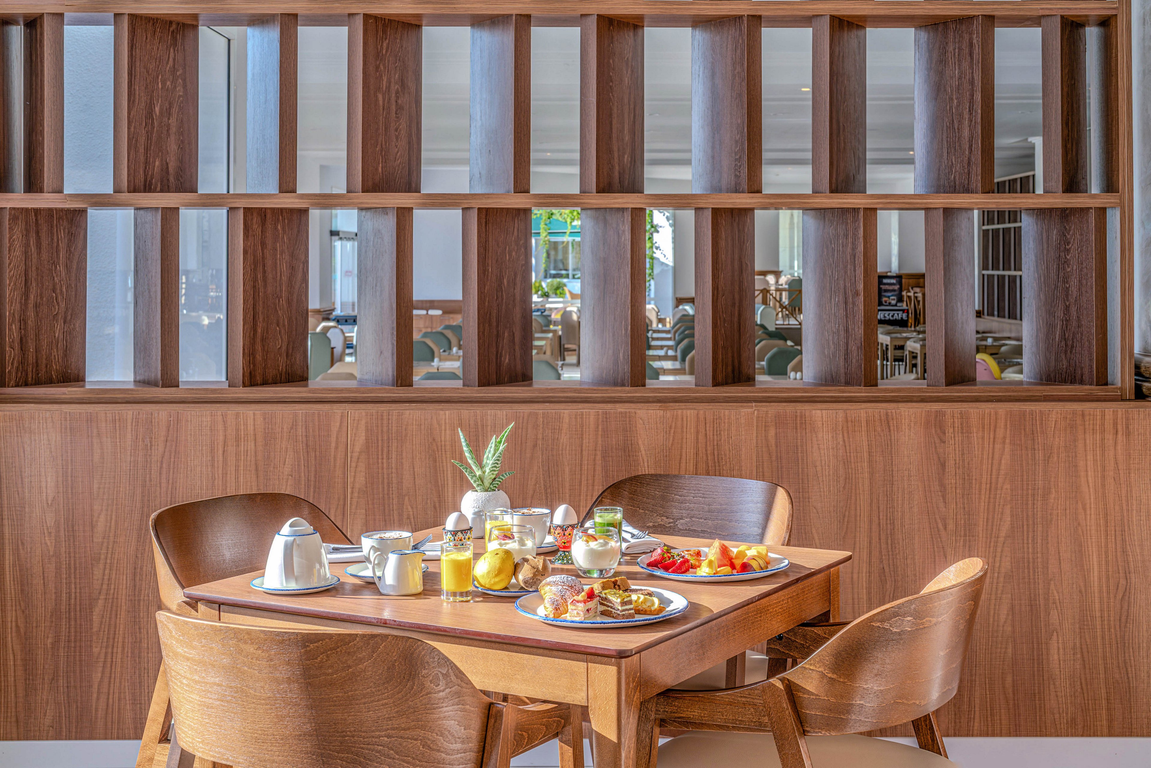 A wooden dining table is set with a generous breakfast spread in a modern hotel restaurant. Plates of pastries, toast, fruit, and eggs are arranged alongside juice, coffee, and condiments. Sleek metal jugs, glassware, and white dishes sit on trays, creating a neat and inviting presentation. In the background, wooden partitions and soft seating suggest a bright, contemporary dining area.
