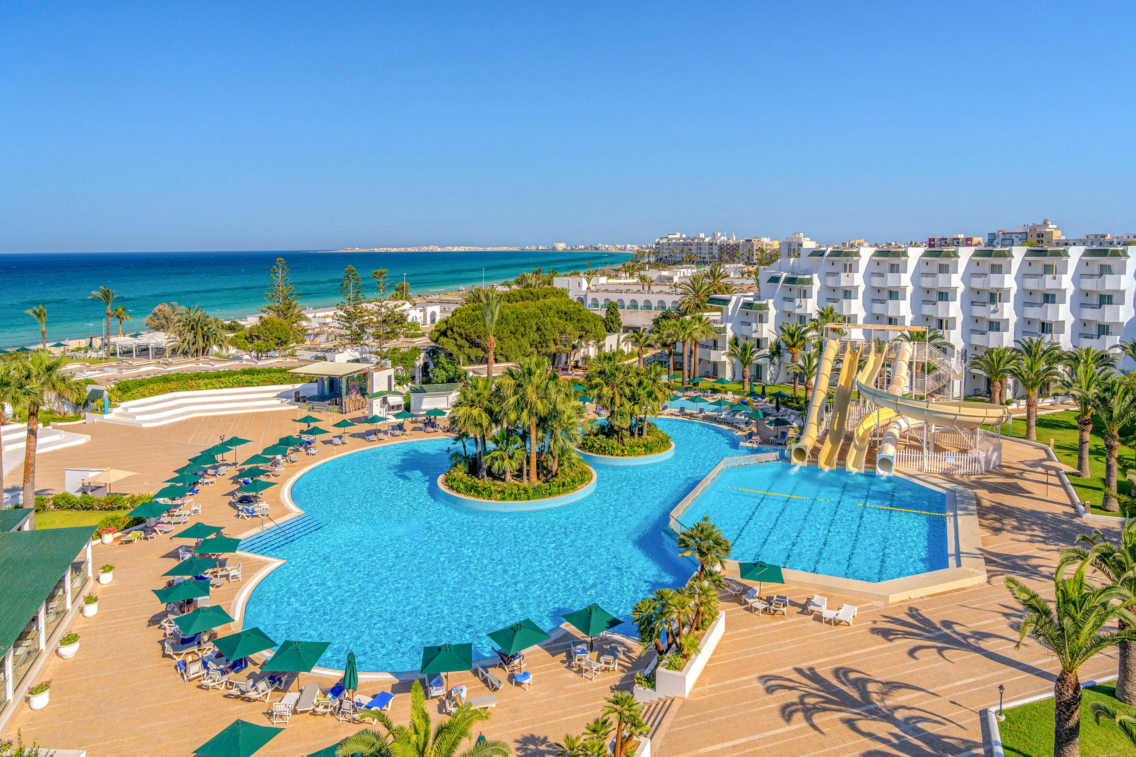 A spacious beachfront resort features a large, irregularly shaped swimming pool surrounded by sun loungers and green umbrellas. Several tall water slides curve into a smaller pool area, adding a sense of fun and activity to the scene. White multi-story hotel buildings line the right side, while palm trees and landscaped gardens create a lush atmosphere. In the background, the turquoise sea and clear blue sky emphasize a sunny, tropical vacation setting.