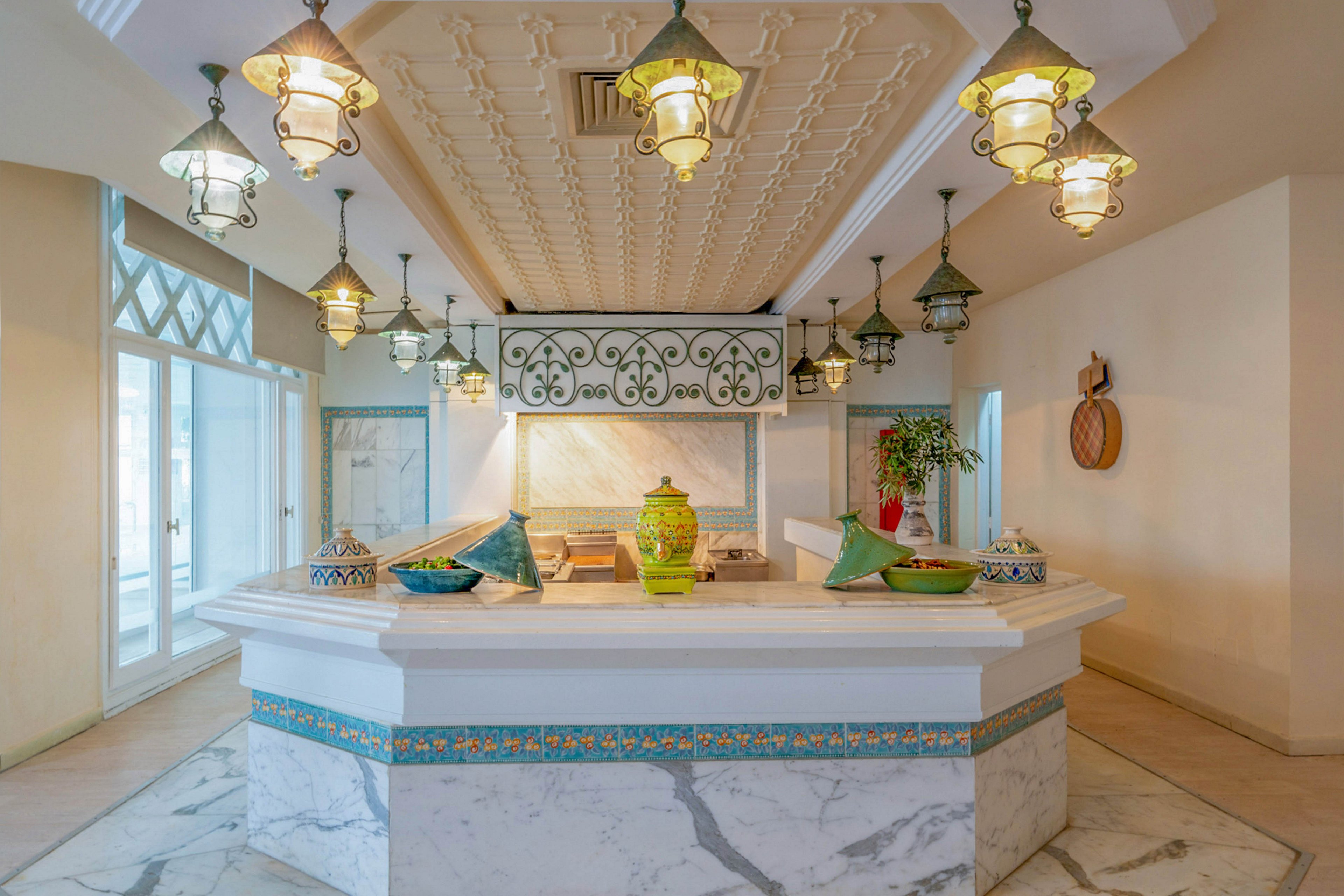 A bright, marble-clad counter stands at the center of an elegant interior space, decorated with colorful ceramic vases and bowls. Ornate hanging lanterns and a patterned ceiling add a Mediterranean or Middle Eastern design touch. Light walls, decorative tiles, and greenery create a welcoming, airy atmosphere. Large glass doors and soft natural light enhance the open, inviting feel of the room.