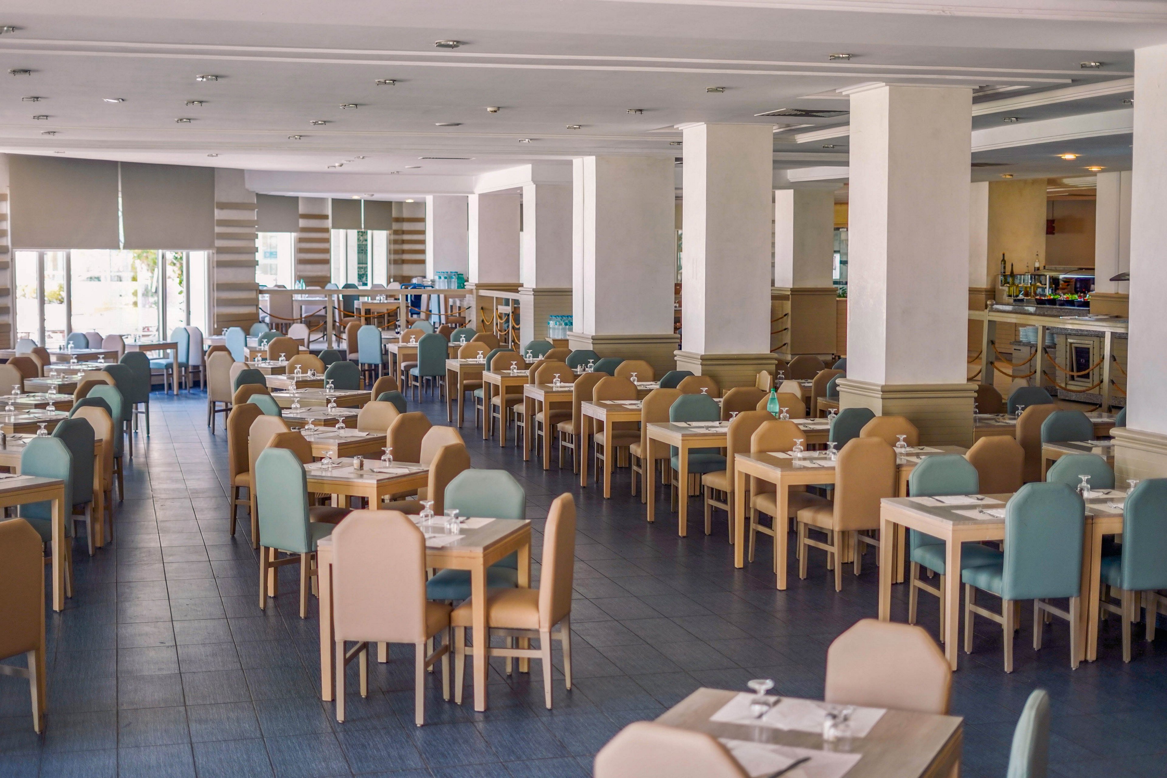 Spacious restaurant dining area with neatly arranged tables and chairs is shown empty and ready for guests. Light wood furniture with pastel blue and beige upholstery creates a soft, modern look. Place settings with glasses and napkins are prepared on each table, and large windows with blinds bring in natural light. The overall atmosphere is clean, orderly, and welcoming, suitable for hotel or resort dining.