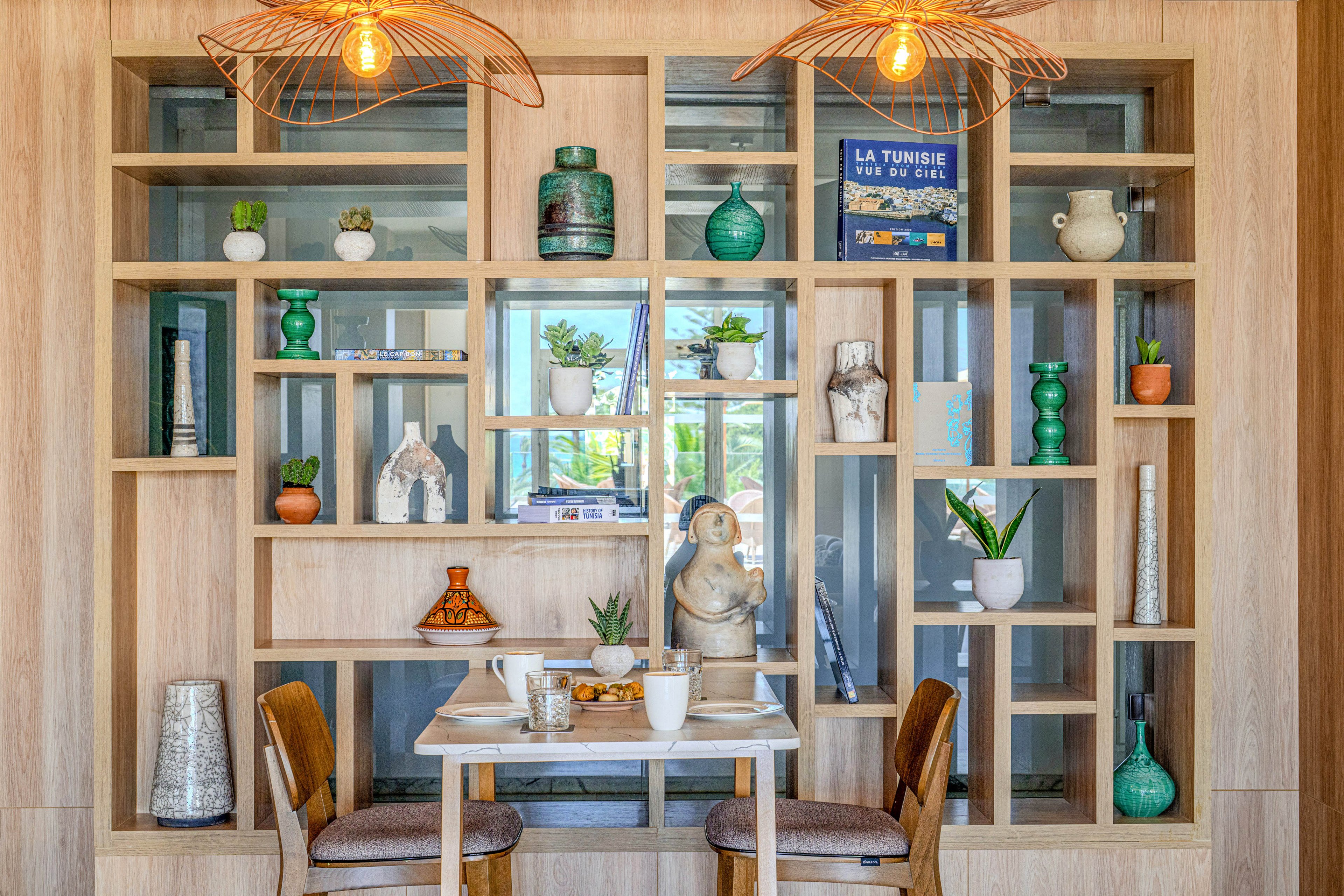 A small dining table set for two is placed in front of a built-in wooden shelving unit. The shelves display various decorative objects including vases, plants, sculptures, and books. Warm pendant lights hang above, casting a soft glow over the scene. A framed poster with the text "LA TUNISIE" is visible on one of the shelves, adding a travel-inspired touch.