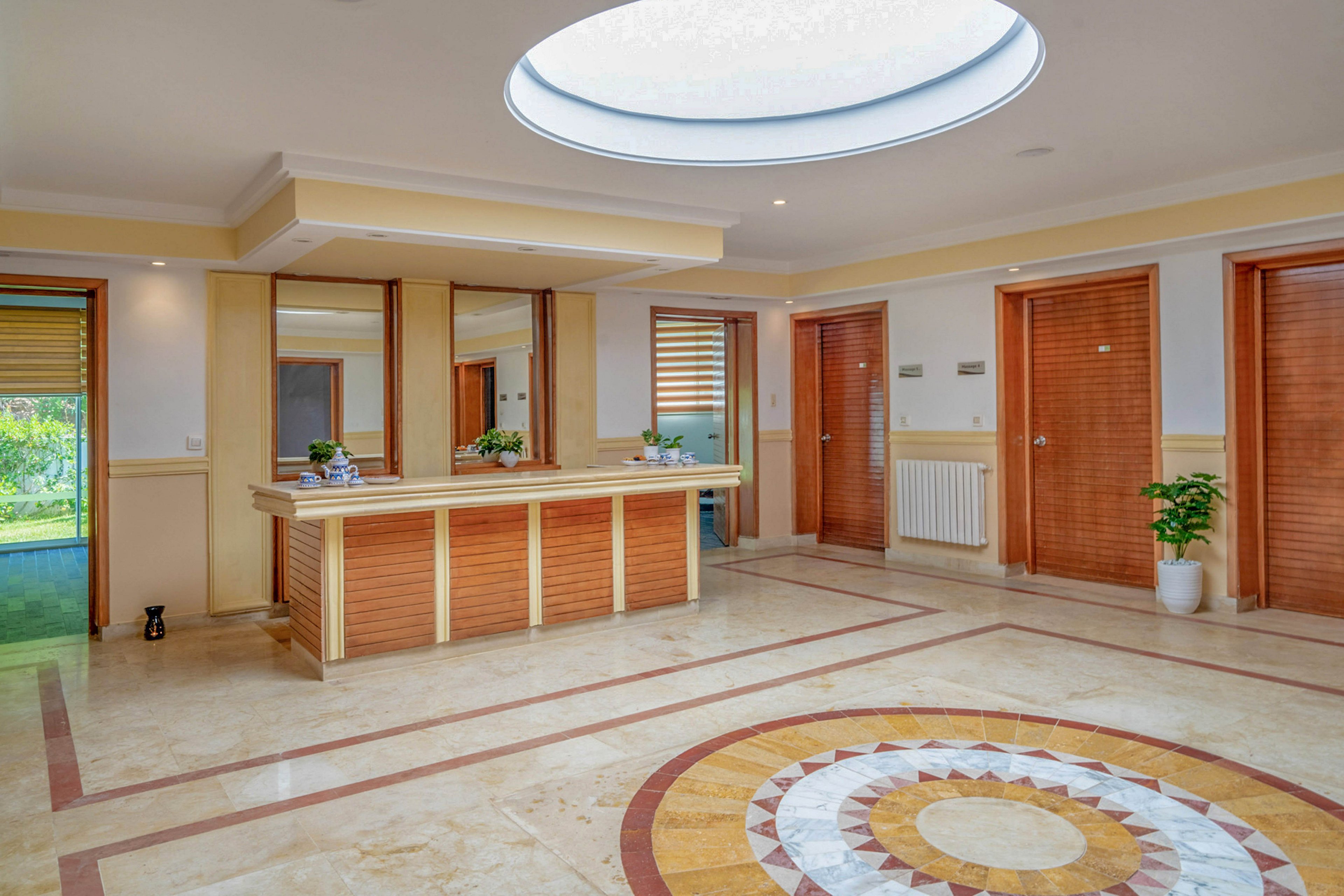 Spacious lobby interior featuring a central reception desk and warm wooden finishes. A large circular skylight illuminates the tiled floor with a decorative round mosaic pattern. Multiple wooden doors and wall mirrors frame the space, with potted plants adding a touch of greenery. Natural light filters in from side windows, creating an inviting and clean atmosphere.
