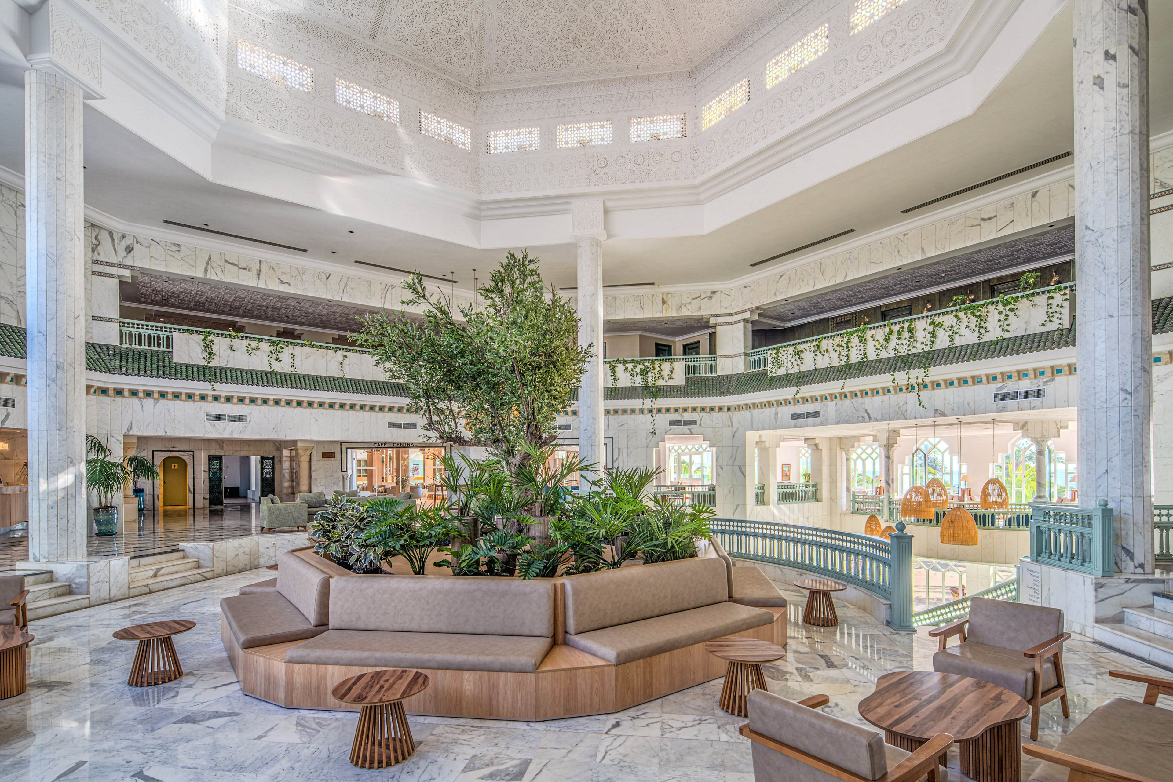 Bright, open atrium interior featuring a large circular seating area surrounding lush indoor plants. The space includes marble floors, white columns, and decorative railings with green accents. Natural light filters through the high ceiling, highlighting multiple levels with arches and balconies. Several wooden tables and chairs create a relaxed lounge atmosphere suitable for socializing or waiting.