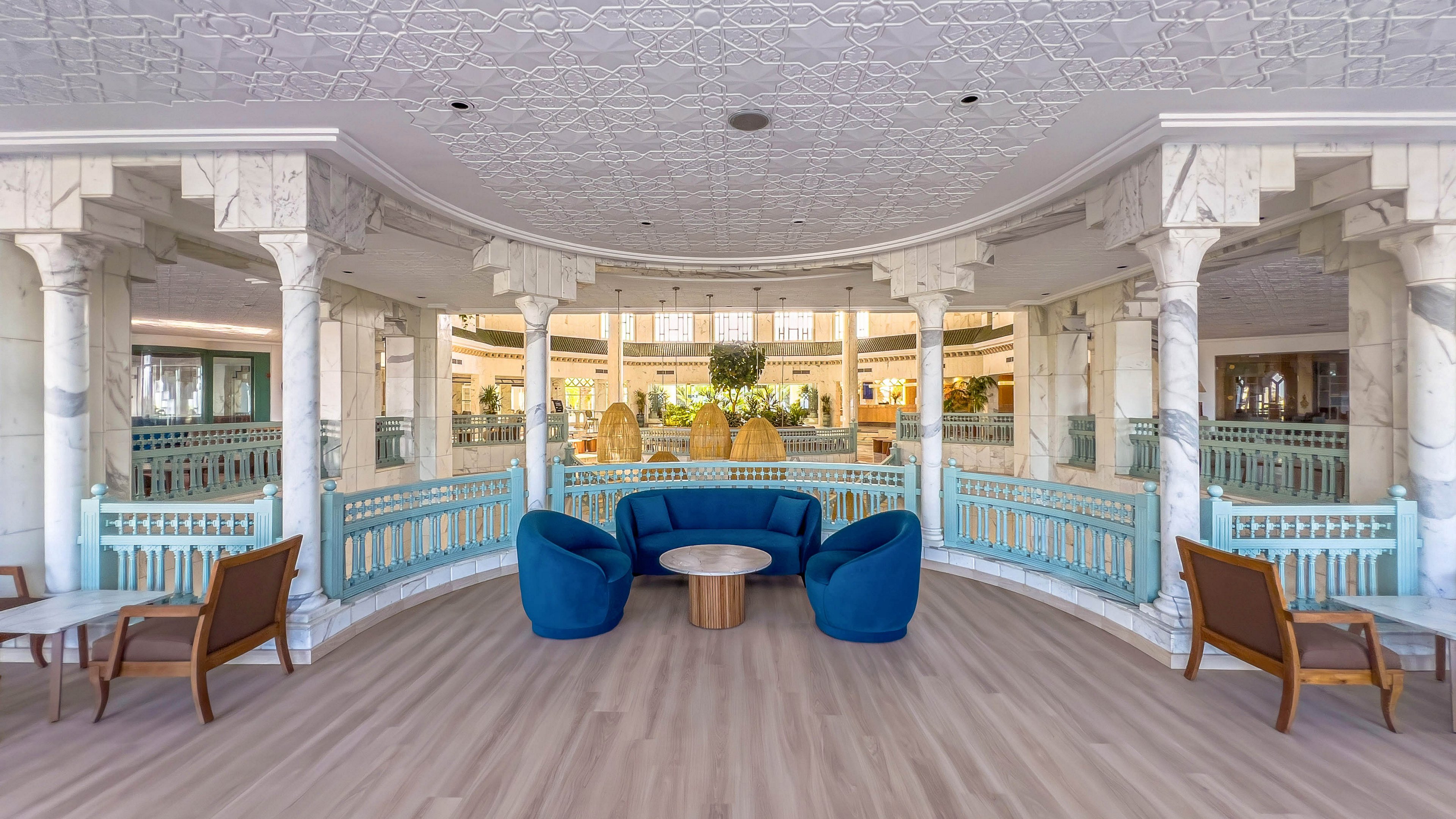 Spacious hotel lobby features a central seating area with modern blue armchairs around a small round table. Ornate white columns and arches frame the space, creating a grand, symmetrical interior. Light wood flooring and pastel railings add warmth and contrast to the cool blue furniture. The background reveals additional seating, greenery, and bright natural light, suggesting a welcoming, upscale environment.