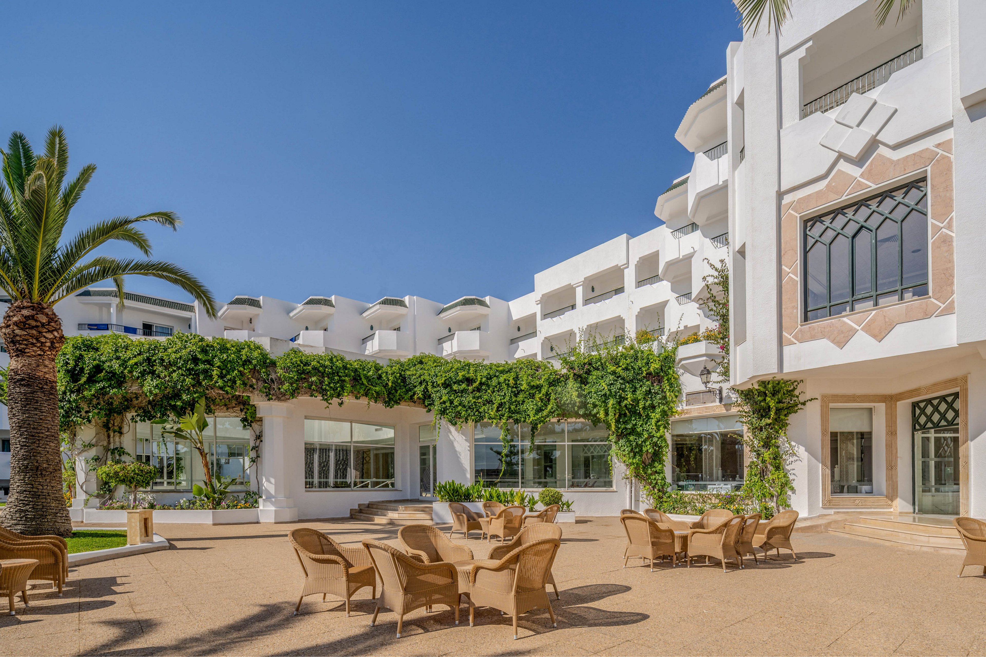 An open-air courtyard of a white Mediterranean-style hotel is shown under a clear blue sky. Wicker chairs and tables are arranged on a spacious terrace, suggesting an outdoor dining or lounge area. Green vines climb along the building façade, adding lush contrast to the bright white architecture. Large windows and balconies overlook the courtyard, creating a relaxed resort atmosphere.