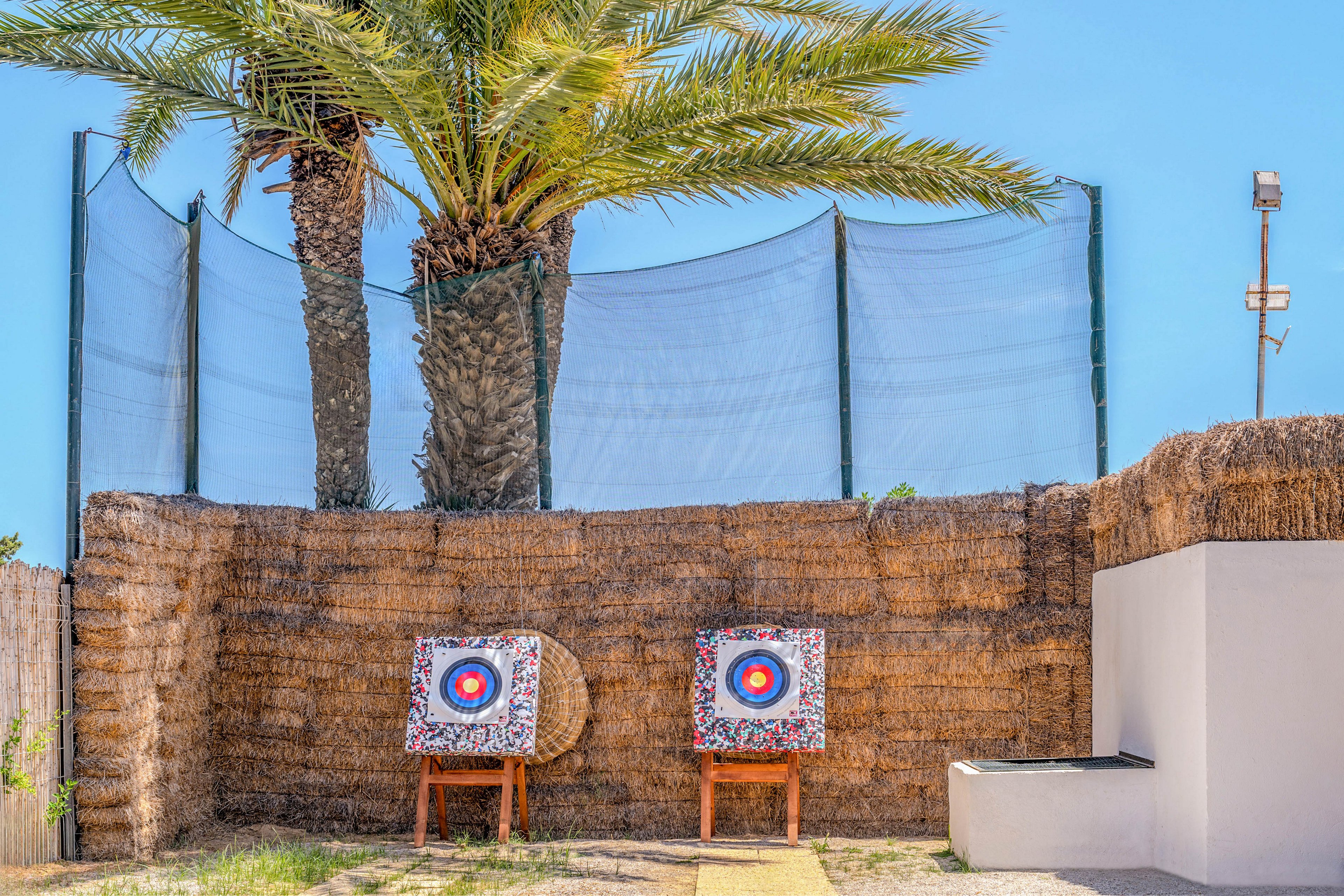 Two colorful archery targets stand on wooden frames in front of a tall straw wall. A palm tree rises behind the wall, with netting stretched above for safety. The scene is set outdoors under a clear blue sky, suggesting a recreational archery range. Bright primary colors on the target faces contrast with the natural tones of straw and greenery.