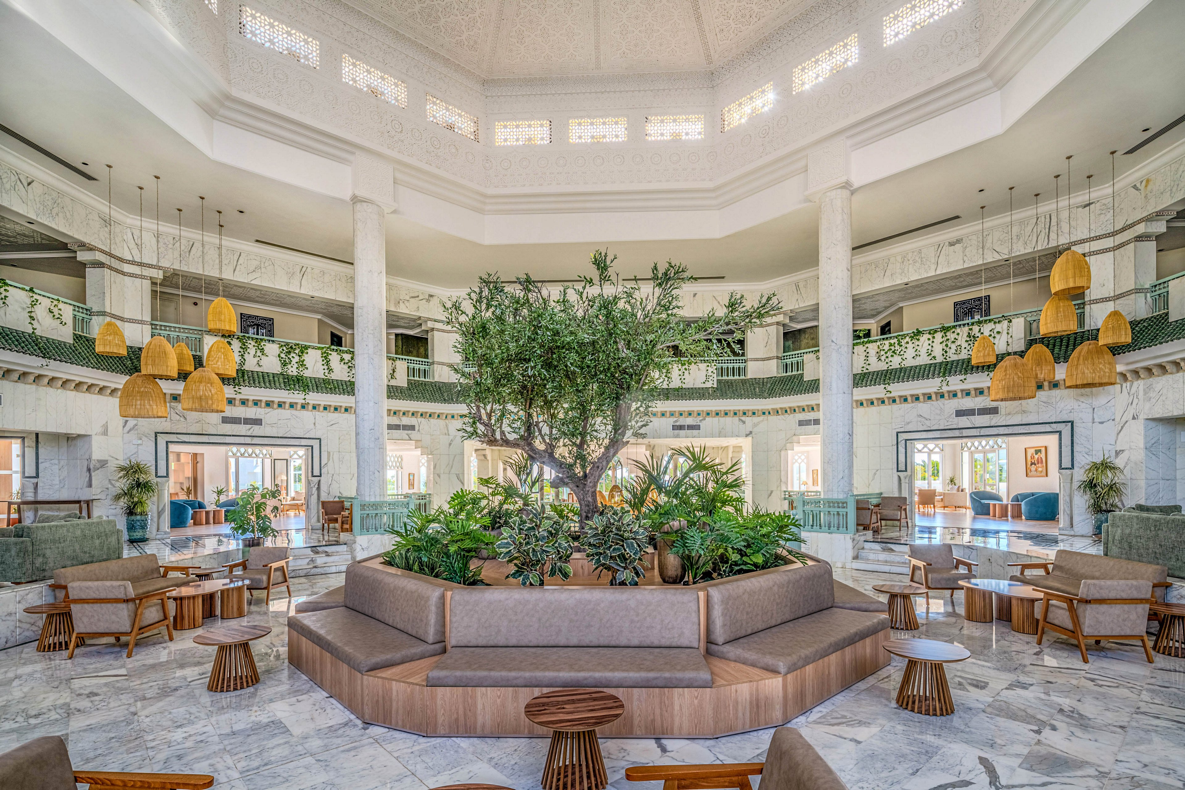 A large, airy hotel lobby features a circular seating area surrounding a lush indoor tree and plants. The space is framed by tall white columns, marble flooring, and decorative railings on the upper level. Warm woven pendant lights hang from the ceiling, complementing wooden chairs and small round tables. Natural light floods the interior, highlighting the modern yet relaxed resort atmosphere.