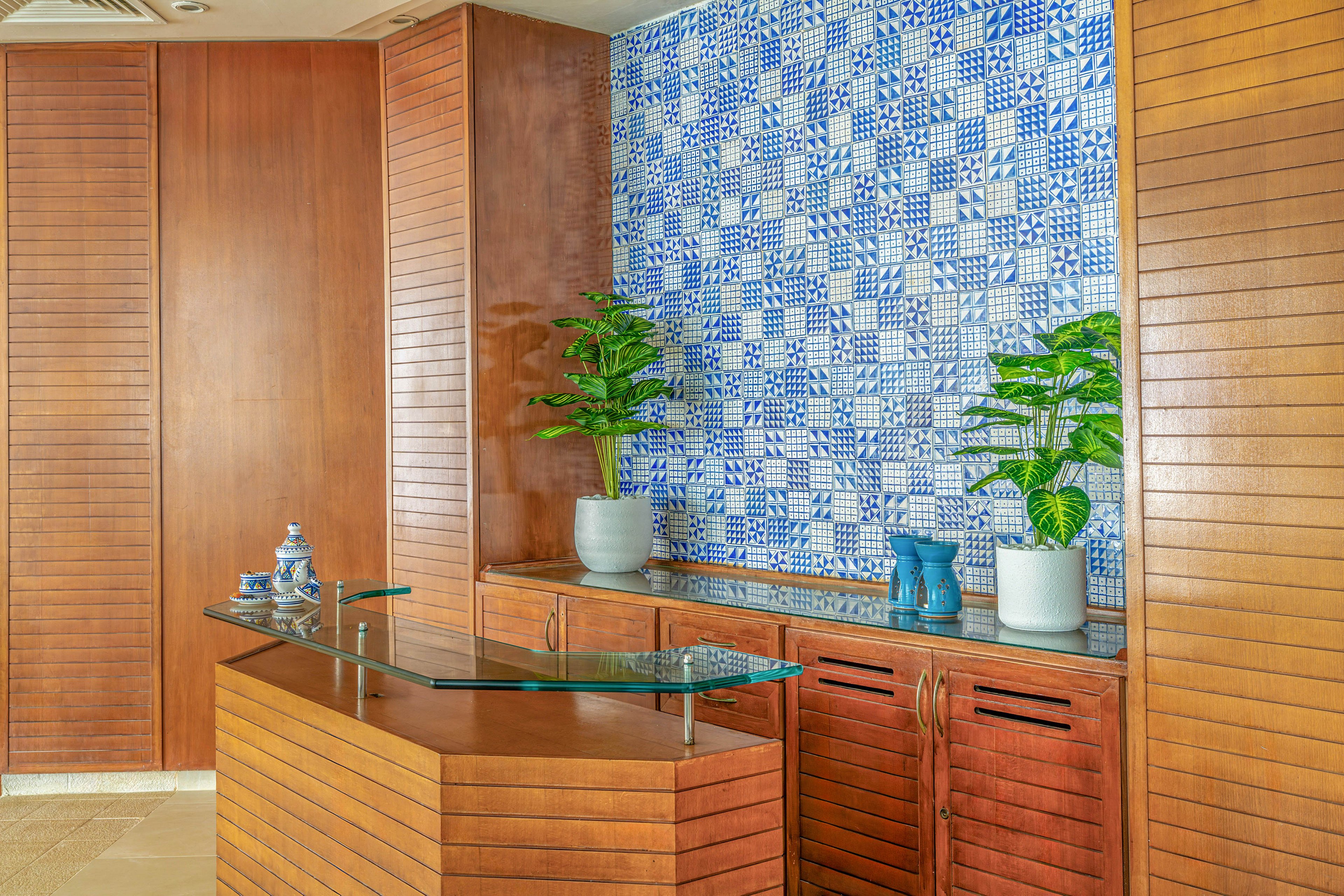A contemporary interior features a wooden reception counter with a glass top in front of a vibrant blue and white patterned tile wall. Two potted green plants sit on the wooden cabinet, adding a touch of nature to the space. Decorative objects, including ceramic pieces and blue vases, are arranged neatly on the surface. The overall design combines warm wood tones with cool blue accents for a clean, modern look.