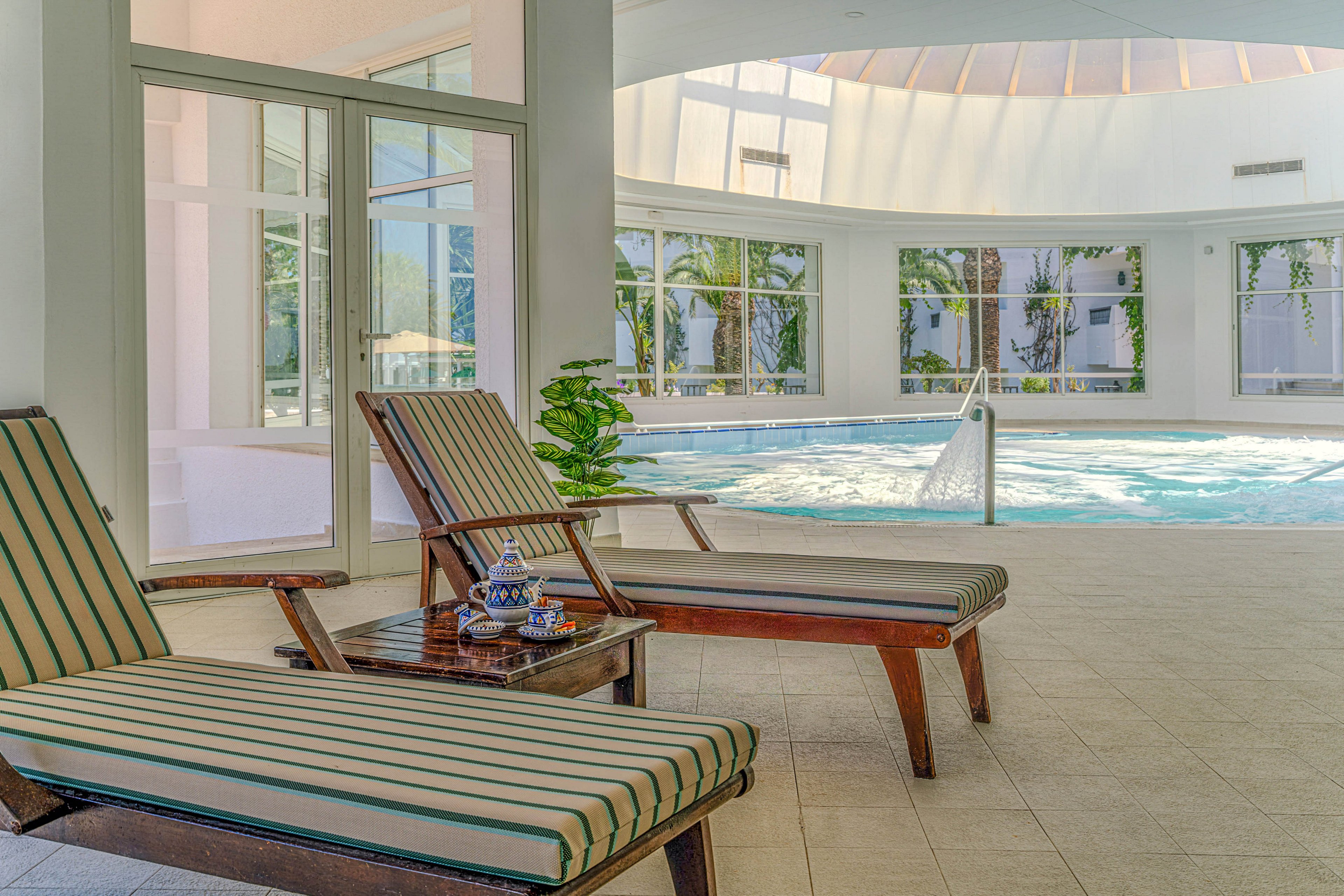 Two striped lounge chairs sit beside a small wooden table in a bright indoor pool area. Sunlight streams through large windows and a curved skylight, illuminating the tiled floor and the water. The pool features a metal handrail and gentle water movement, suggesting a relaxing spa-like environment. Green plants outside the windows add a touch of nature to the serene setting.