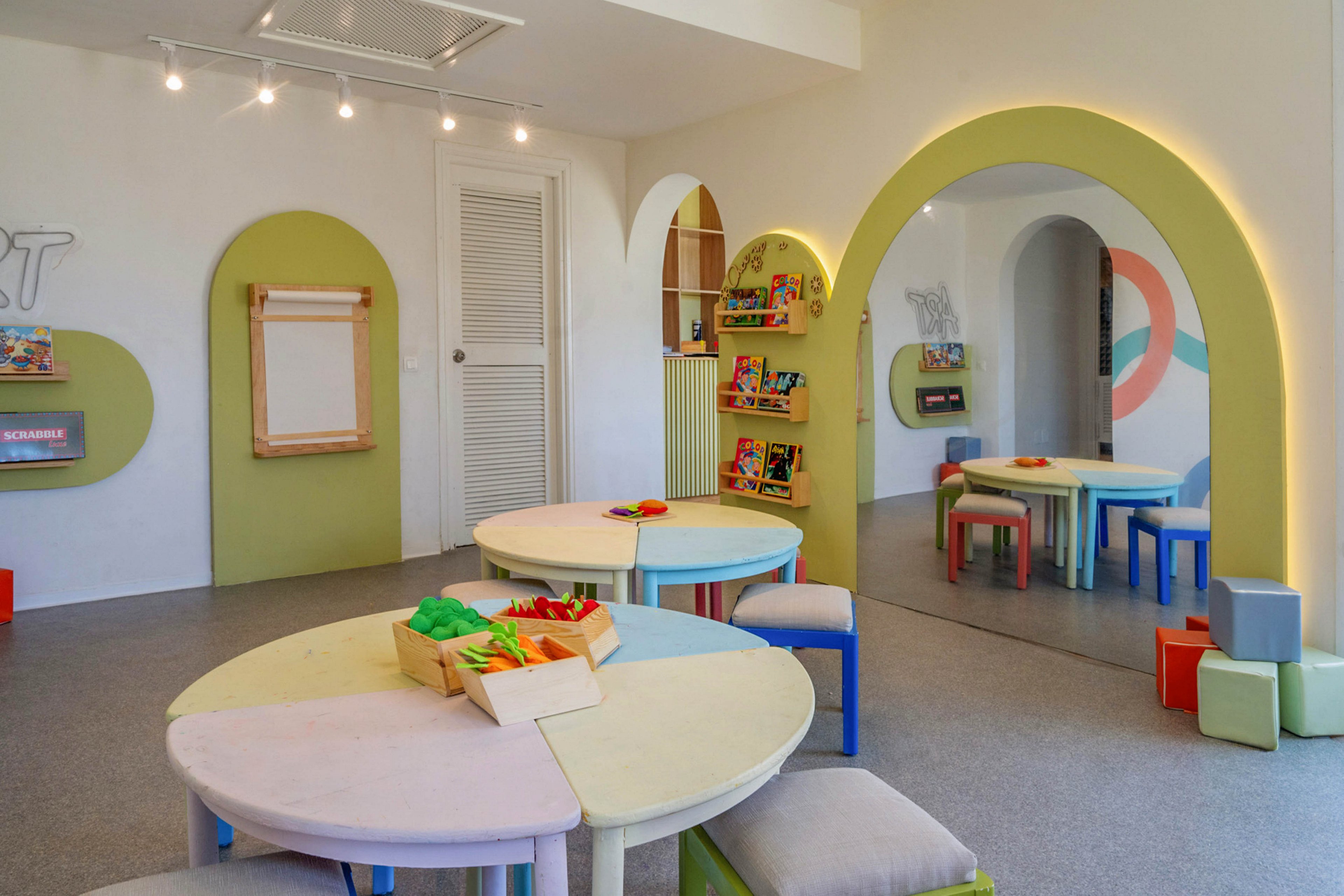 Bright, contemporary children's playroom featuring round tables and small stools in pastel and primary colors. Wooden trays with toys and building blocks sit on the tabletops, inviting creative play. Curved wall openings, built-in shelves with books and games, and a large wall mirror enhance the playful design. Soft lighting and rounded shapes create a welcoming, safe environment for kids' activities.