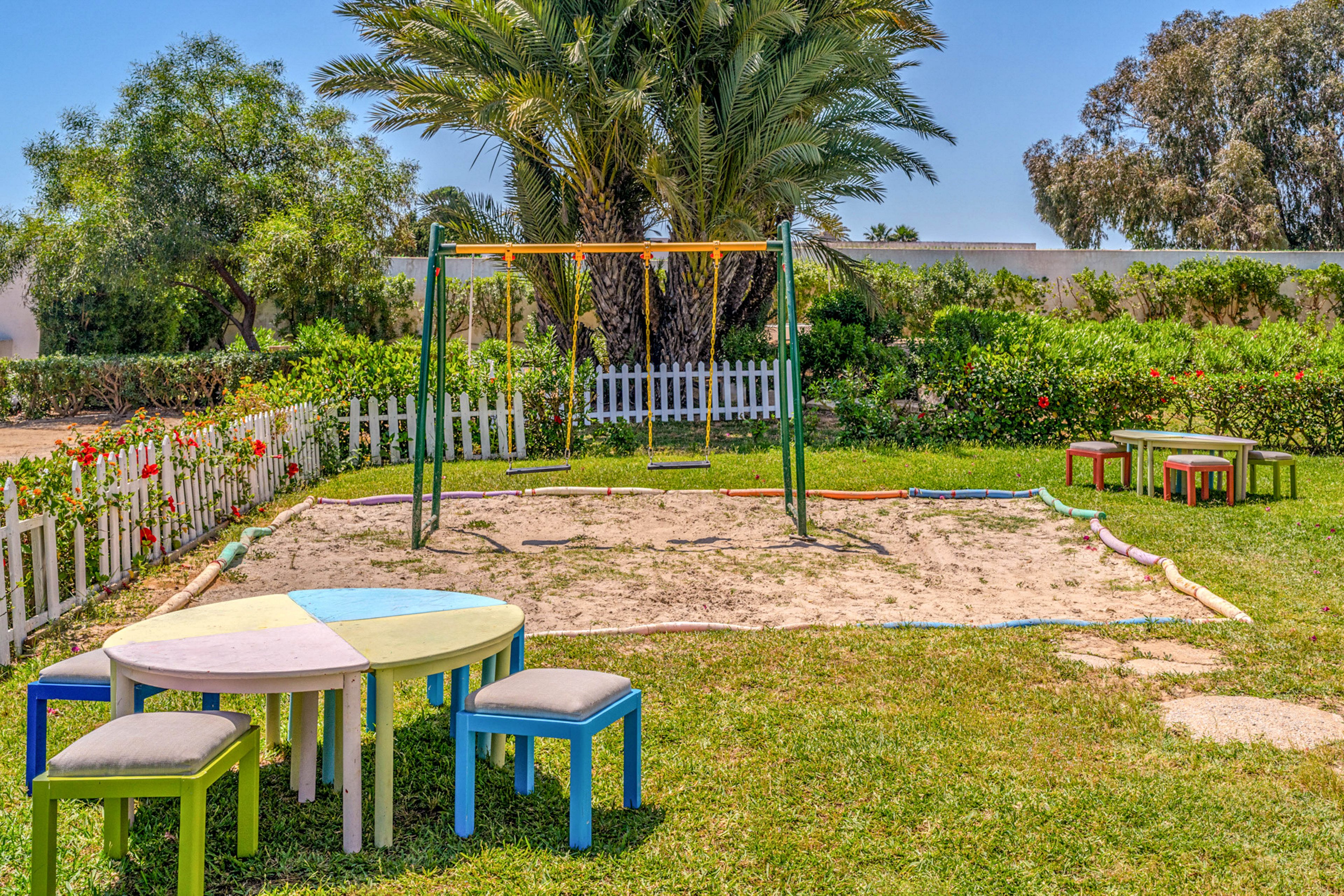 An outdoor playground features a sandy area with a metal swing set at its center. In the foreground, round and rectangular colorful tables with matching stools sit on the grass. The play area is surrounded by a white picket fence, flowering bushes, and lush green trees and palms. Bright daylight and clear skies create a cheerful, inviting atmosphere for children.