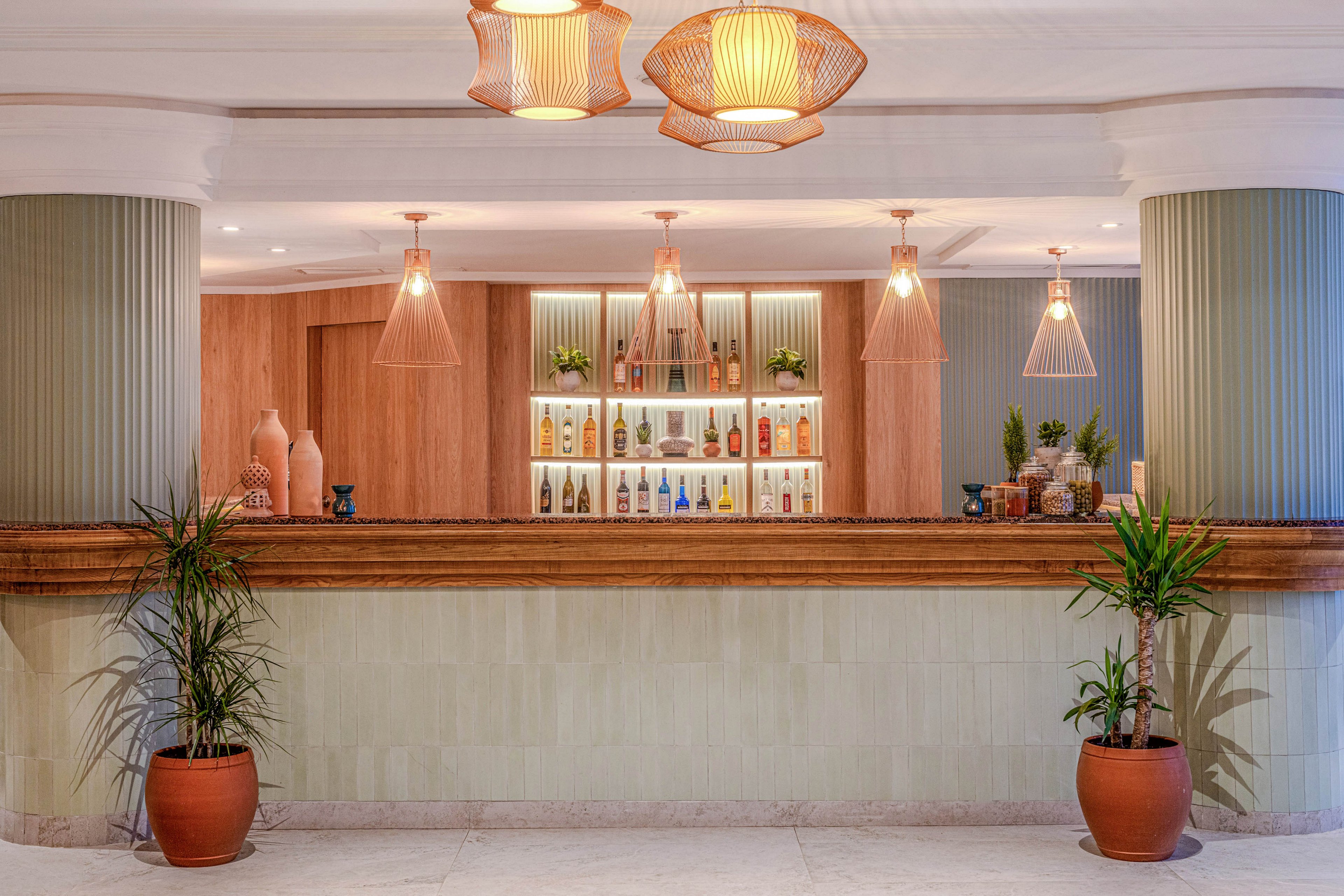 A contemporary bar counter is shown with a clean, minimalist design and warm wooden accents. Backlit shelves display an assortment of colorful bottles and glassware neatly arranged. Pendant lights with soft, warm bulbs hang from the ceiling, casting a cozy glow over the scene. Large potted plants frame the bar area, adding a touch of greenery to the sophisticated interior.
