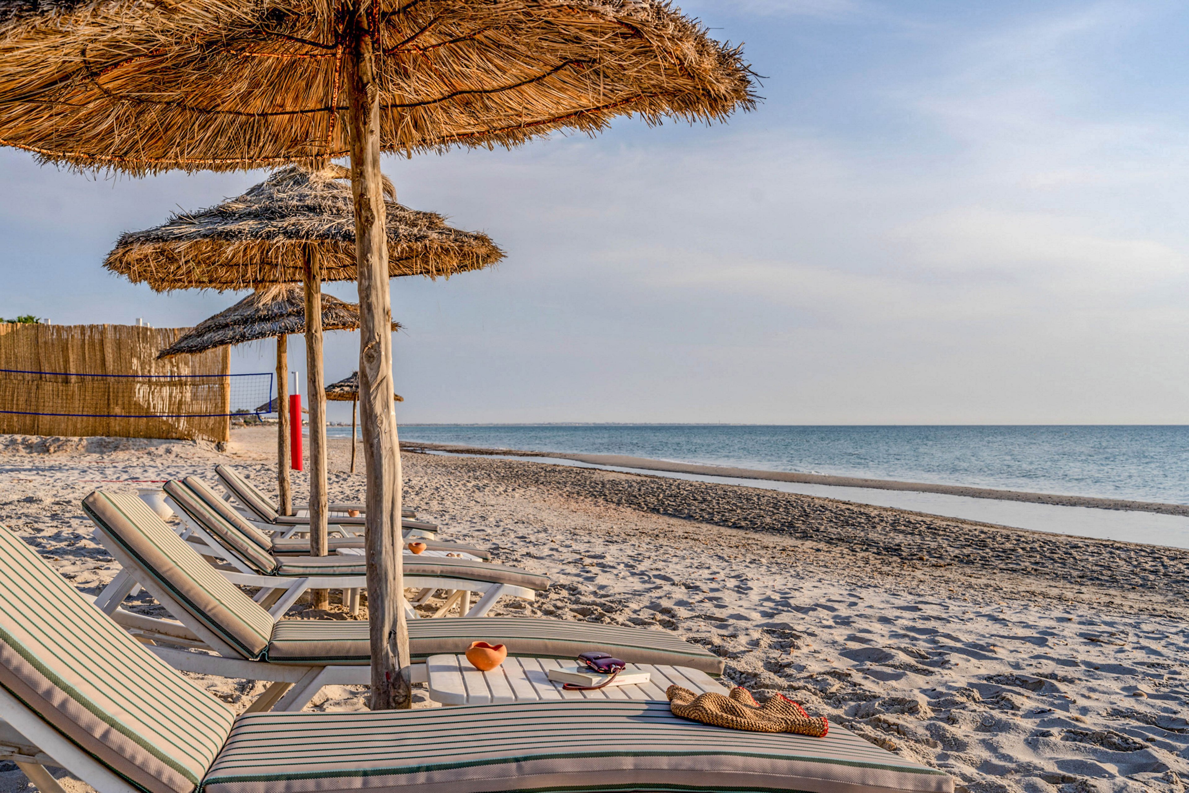 A row of cushioned sun loungers sits on a sandy beach beneath large straw umbrellas. The shoreline stretches into the distance beside calm, blue sea water under a soft, hazy sky. A wooden fence and a red flotation device are visible in the background, adding detail to the coastal setting. The scene feels peaceful and inviting, suggesting a quiet seaside resort at sunset or early evening.