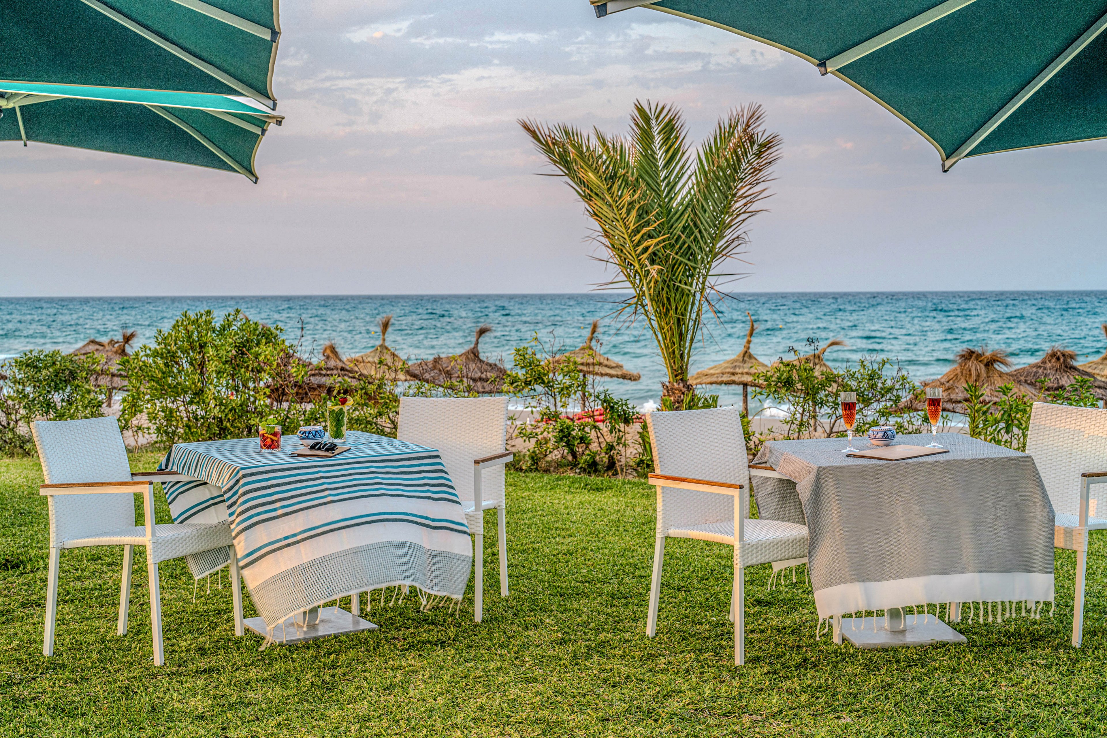 An inviting outdoor dining setup is arranged on a grassy lawn overlooking the sea. White chairs surround tables draped with striped and solid throws, with drinks and tableware placed neatly on top. Large teal umbrellas provide shade, while straw parasols line the beach in the background. The calm ocean and soft sky create a relaxed, vacation-like atmosphere.