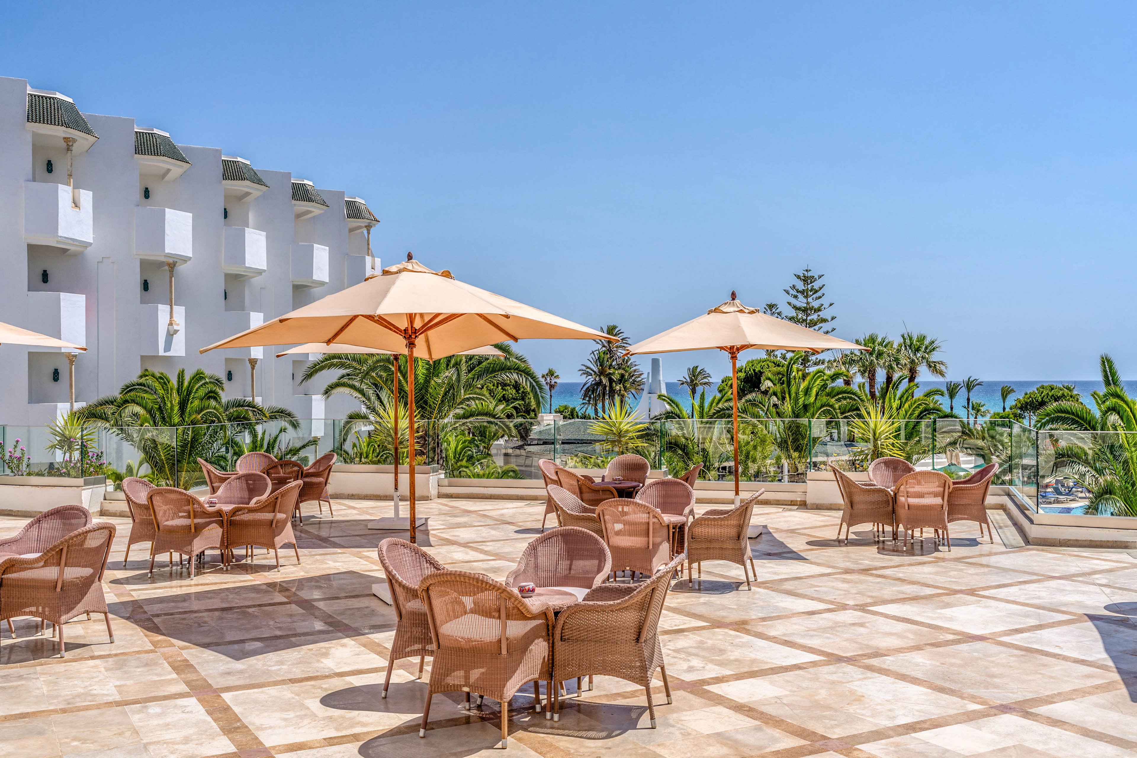 An open-air resort terrace features wicker chairs and tables arranged beneath large beige umbrellas. The setting overlooks lush palm trees and a distant view of the sea under a clear blue sky. A white multi-story building with balconies lines one side of the terrace, adding to the vacation atmosphere. The bright sunlight and clean tiled floor create a relaxed, inviting space for guests to sit and enjoy the coastal scenery.